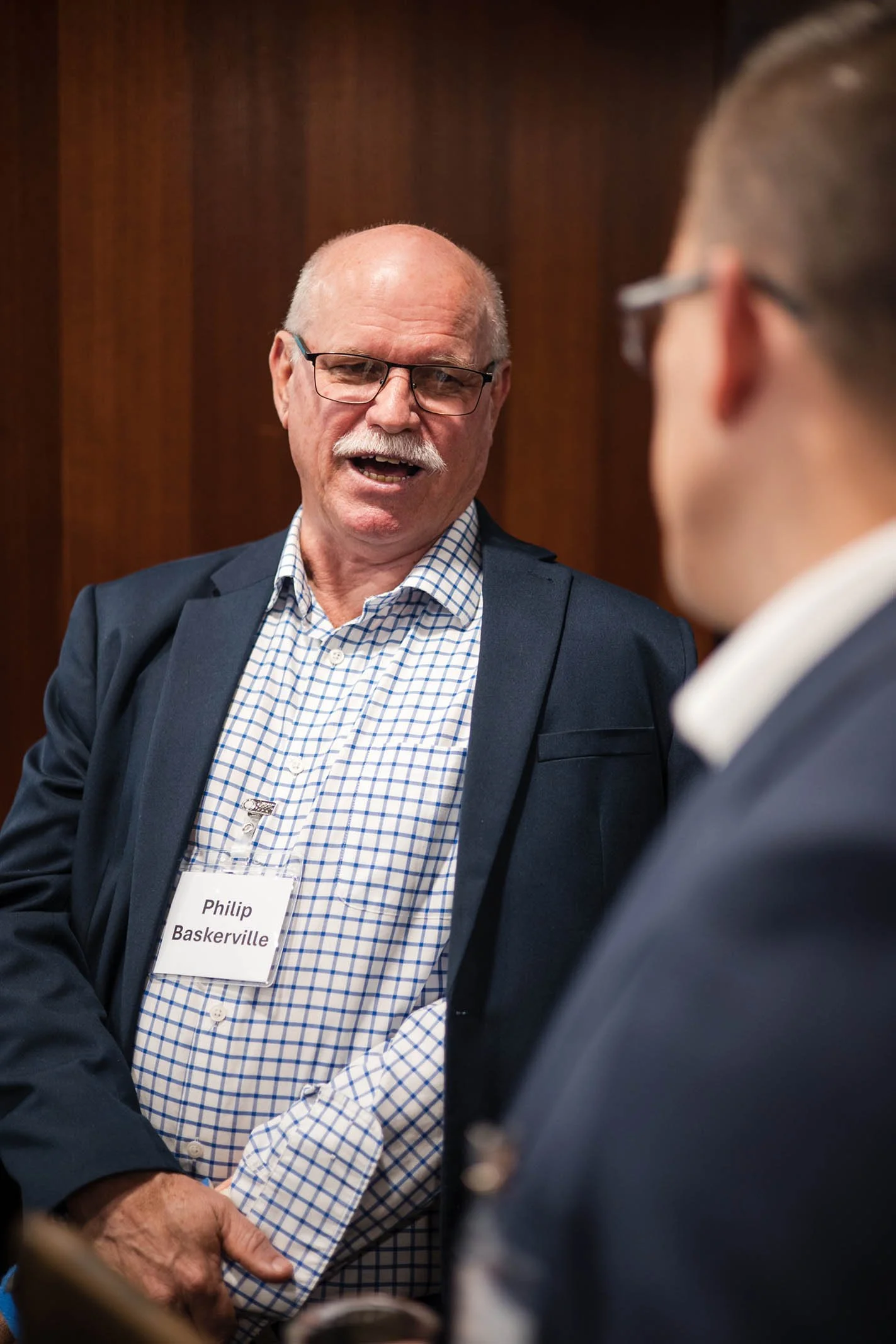 An older man with glasses, a white mustache, wearing a checkered shirt and a dark blazer, is speaking to a younger man with glasses in a formal setting. The older man has a name tag that reads 'Philip Baskerville'.