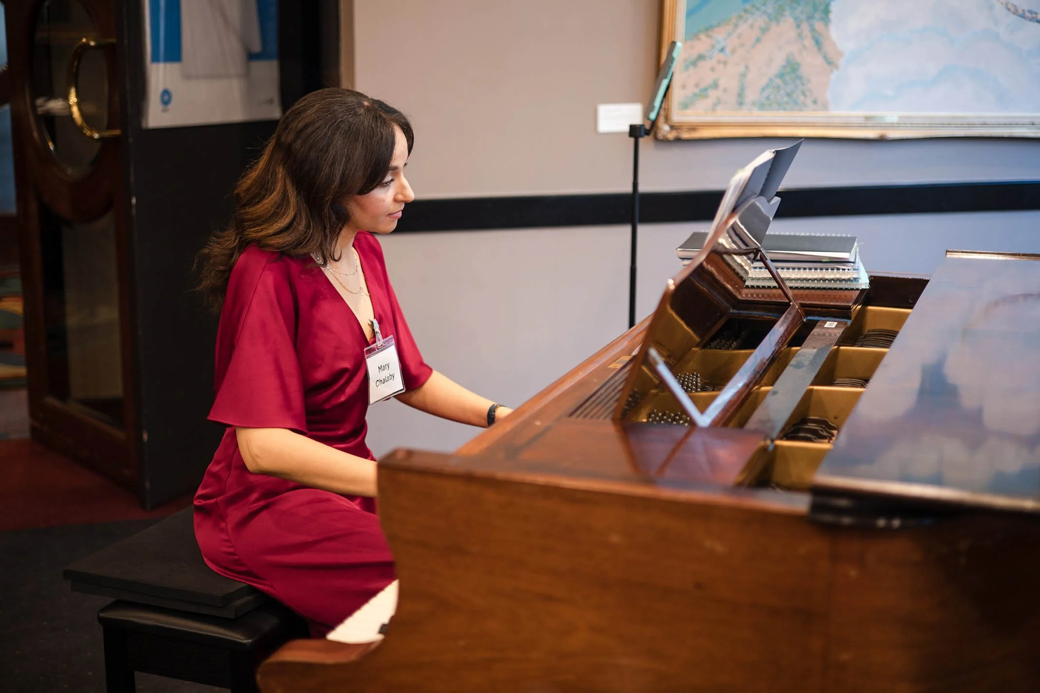 A woman wearing a maroon dress playing a grand piano in a room with artwork on the wall.