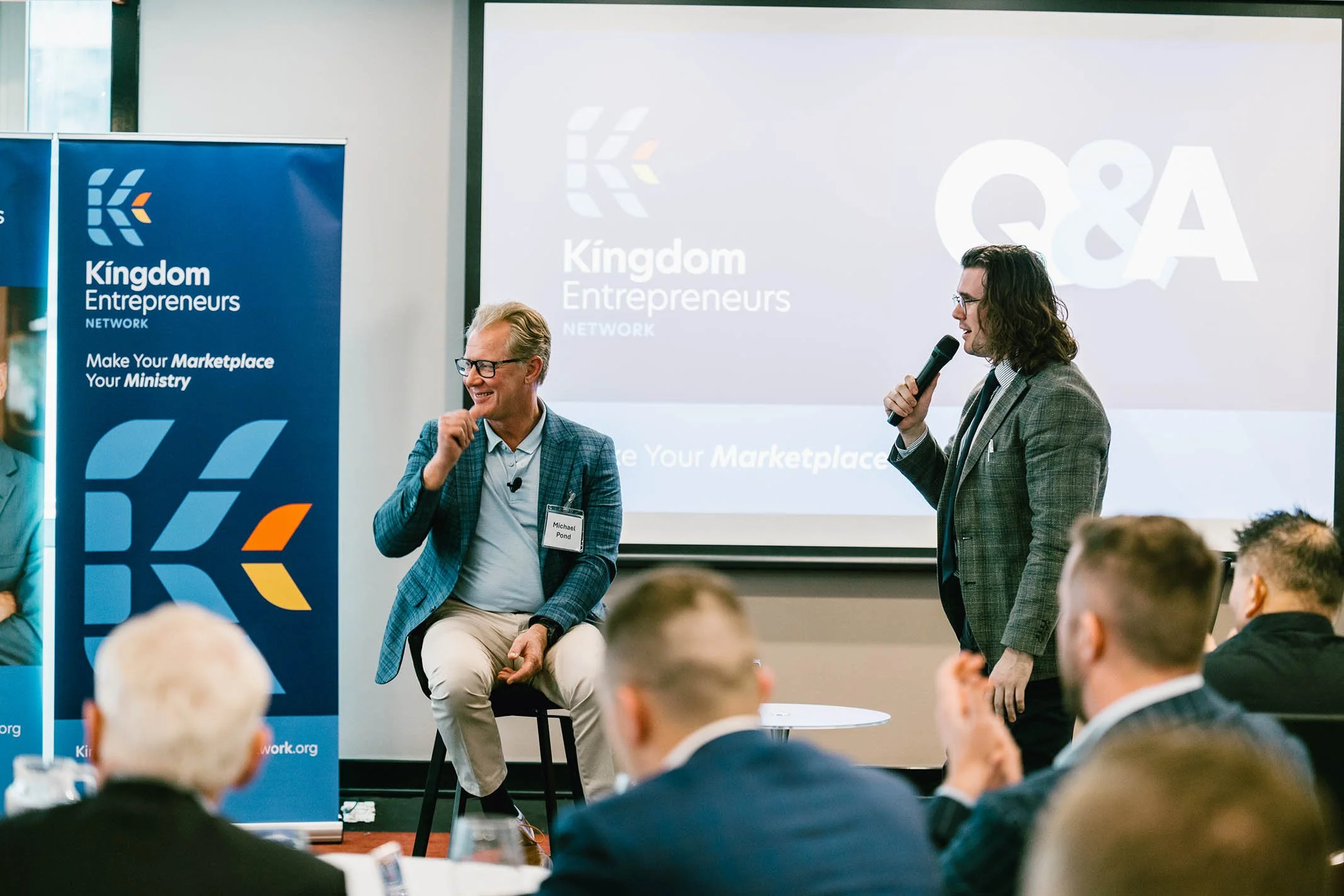Two men speaking at a conference, one is sitting on a stool with a name tag that reads Michael Pond, and the other is standing with a microphone. They are behind a banner for Kingdom Entrepreneurs Network with a blue background and logo and a present