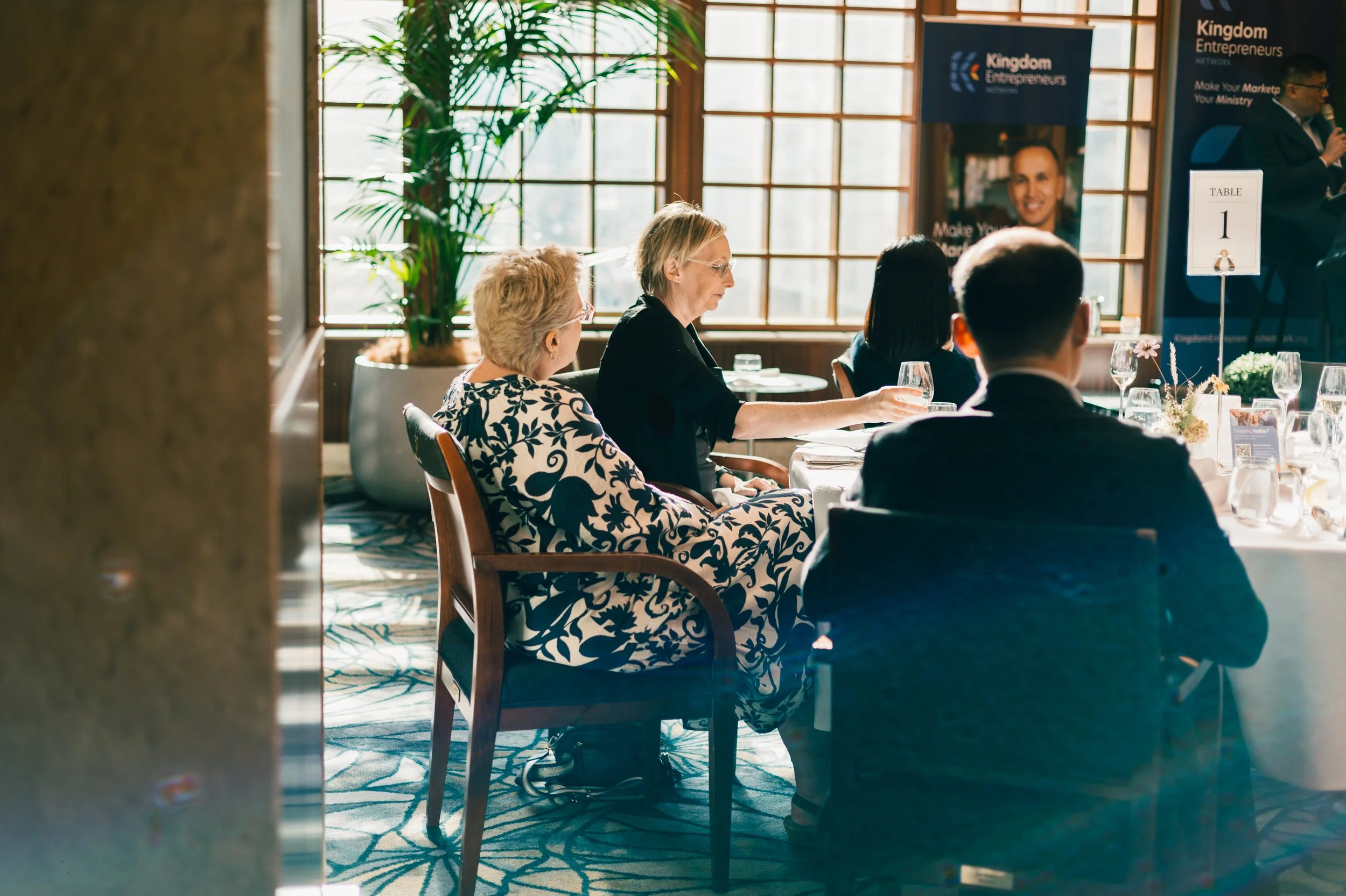 People seated at a banquet table during a formal event with a speaker on stage, banners, and table number 1 visible.