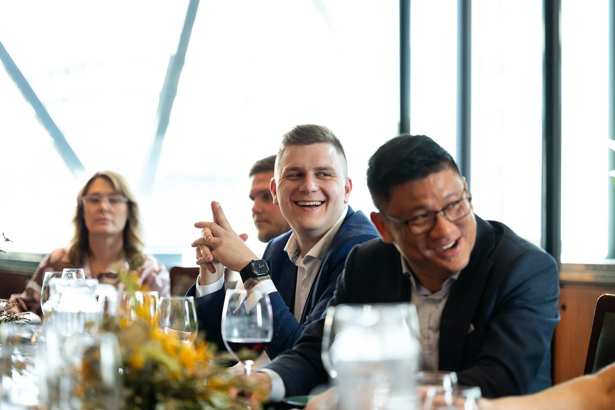 Group of diverse professionals sitting at a conference table during a meeting, smiling and engaging in discussion, with wine glasses and a floral centerpiece on the table.