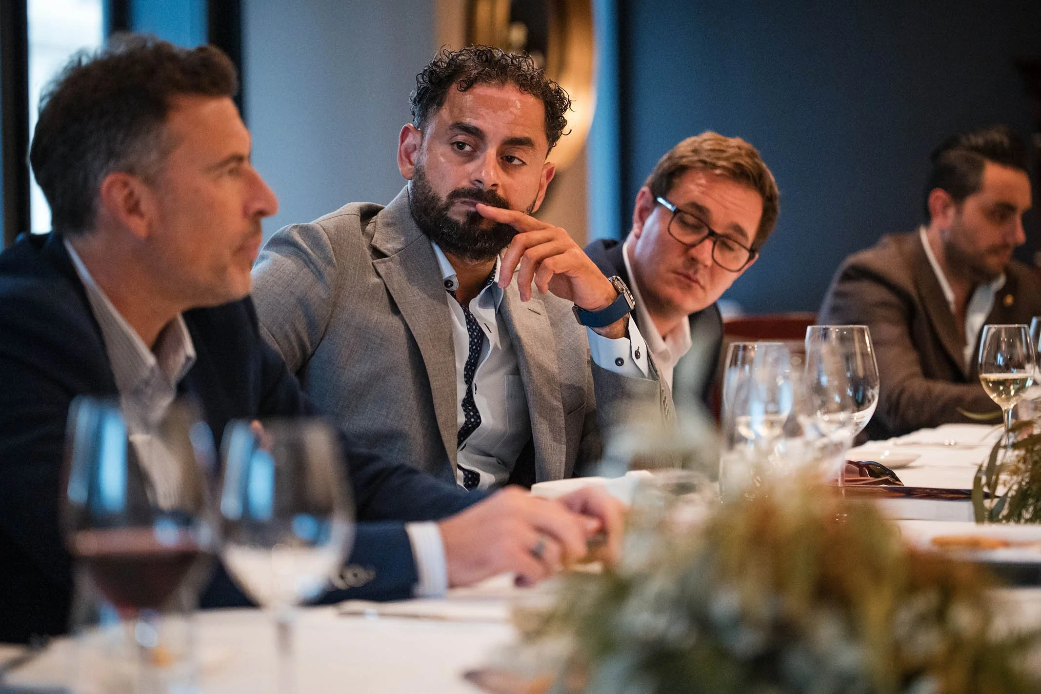 Businessmen sitting at a conference table during a meeting, with glasses of water and wine in front of them, in a formal setting.