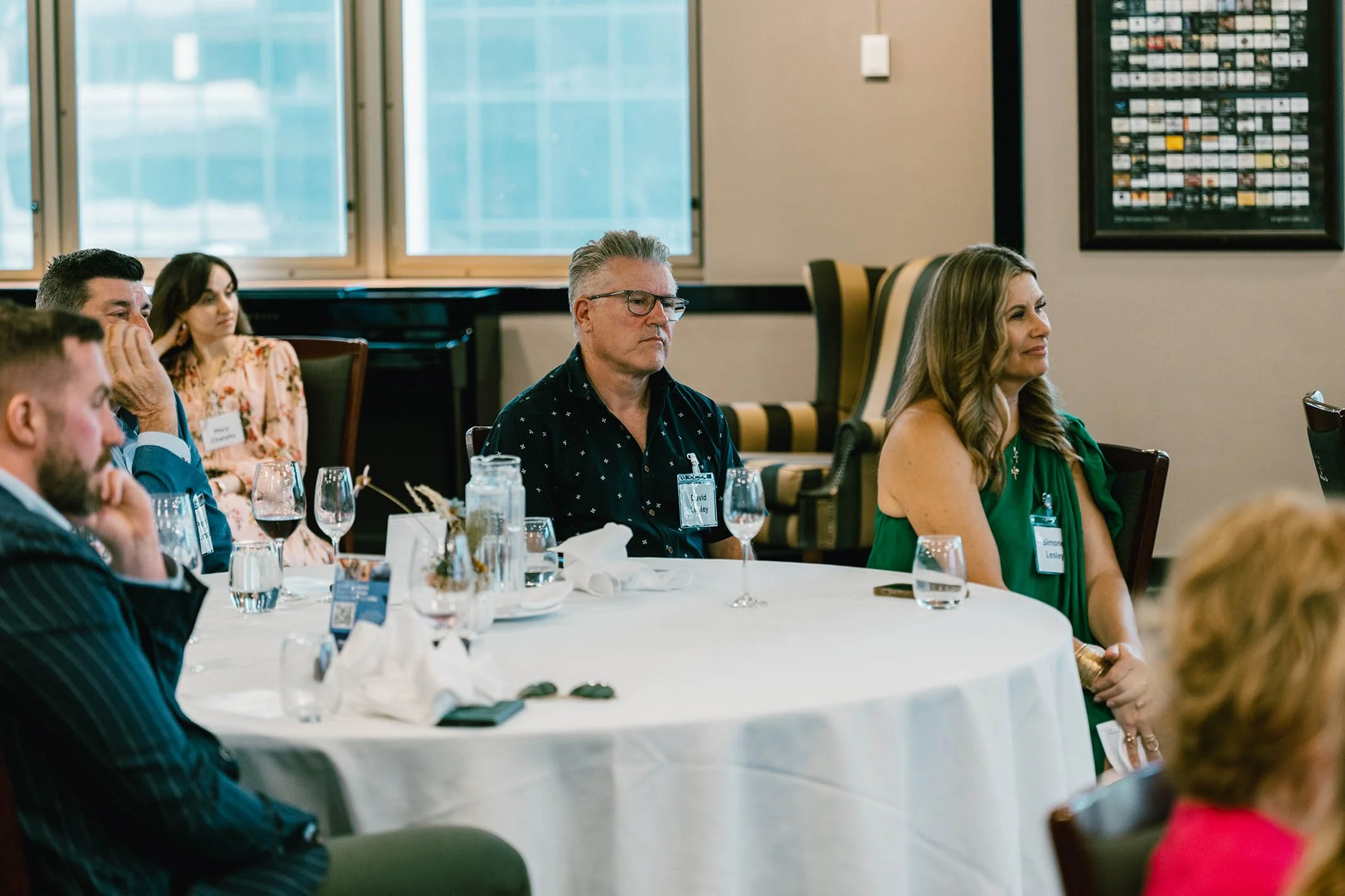 People sitting at a round table during an indoor conference or meeting, with glasses and bottles of water on the table.