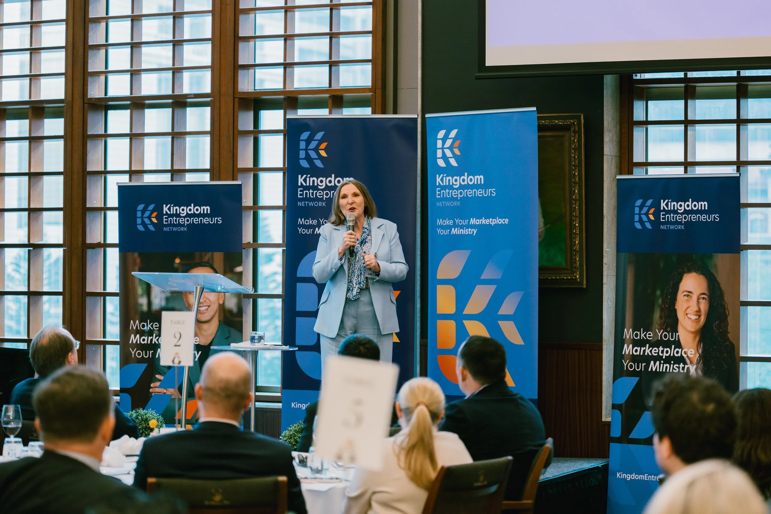 A woman in a light blue suit speaking into a microphone at a Kingdom Entrepreneurs event, with audience members sitting at tables in front of her, and multiple blue banners with the organization's logo and slogan behind her.