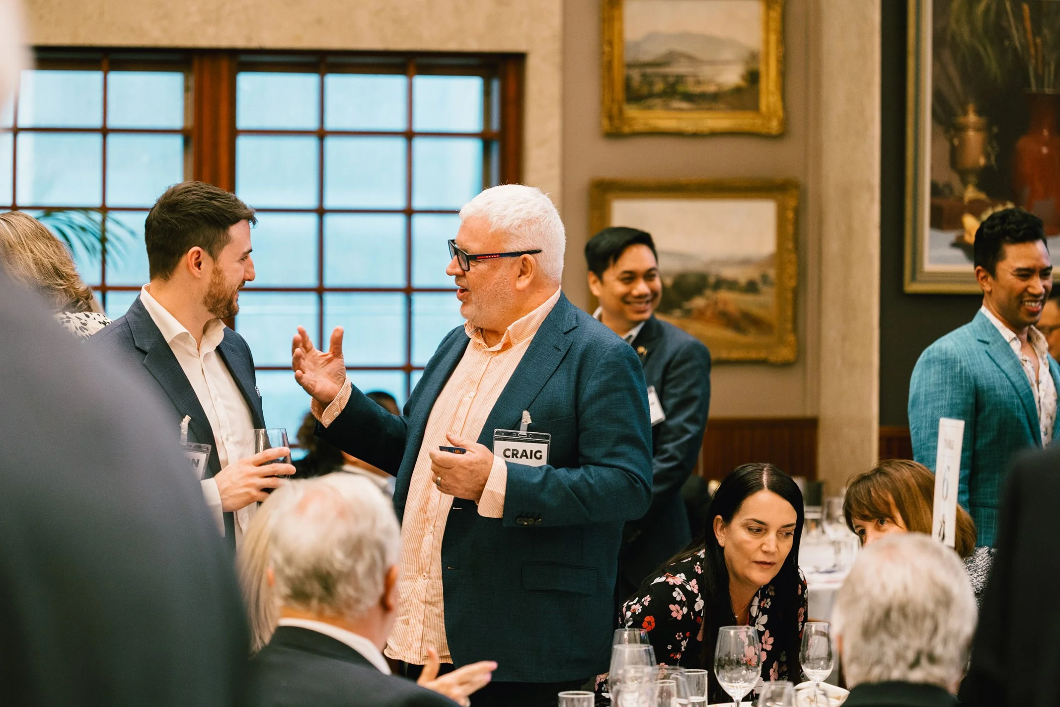 A group of people at a formal event or conference, engaging in conversation. In the center, an older man with white hair, glasses, and a name tag reading 'CRAIG' is speaking to a younger man holding a glass of wine. Other attendees are seated at tabl