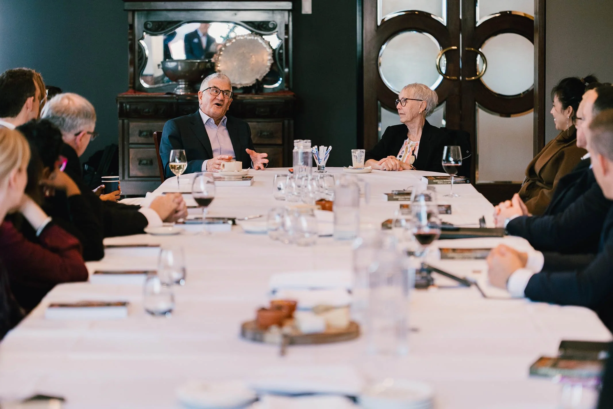 A formal business meeting with people sitting around a long table, listening to two speakers at the head. The man on the left is gesturing while talking, and the woman next to him is listening attentively. The table has glasses, notebooks, and snacks