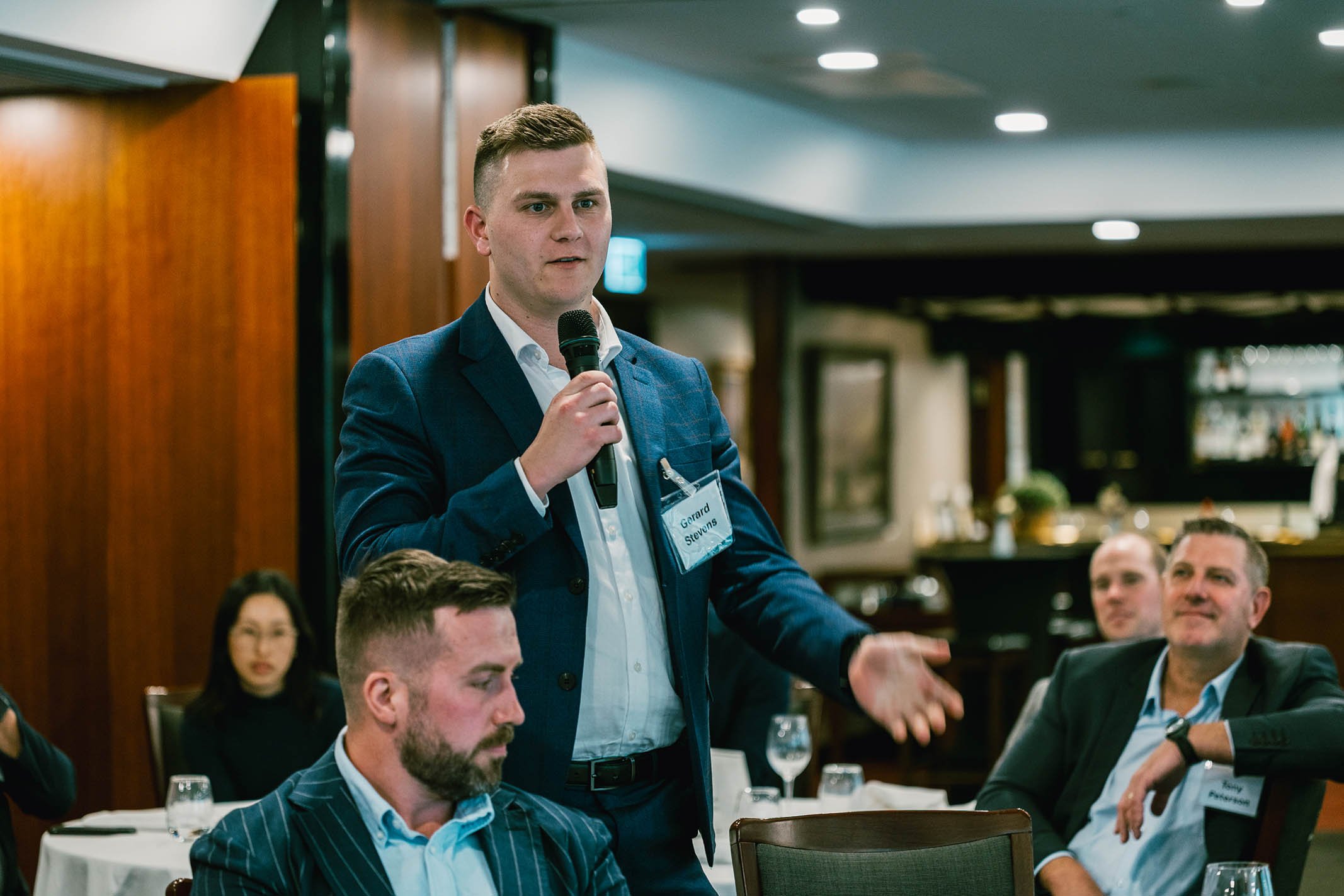 A young man in a blue suit speaking into a microphone during a business meeting or conference, seated at a table with other attendees, some of whom are listening attentively.