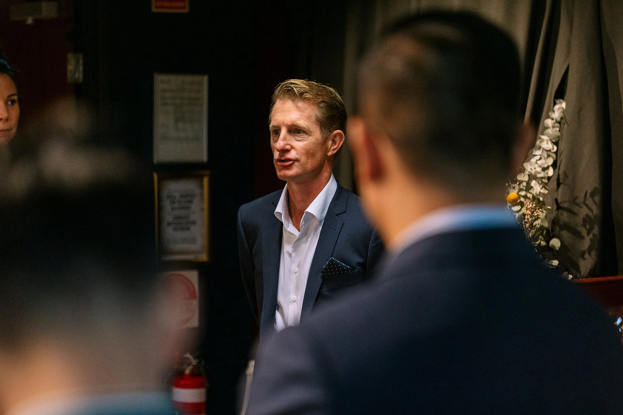 A man in a navy suit and white shirt speaking to a group in a dimly lit room, with a woman partially visible on the left and a blurred person in the foreground.