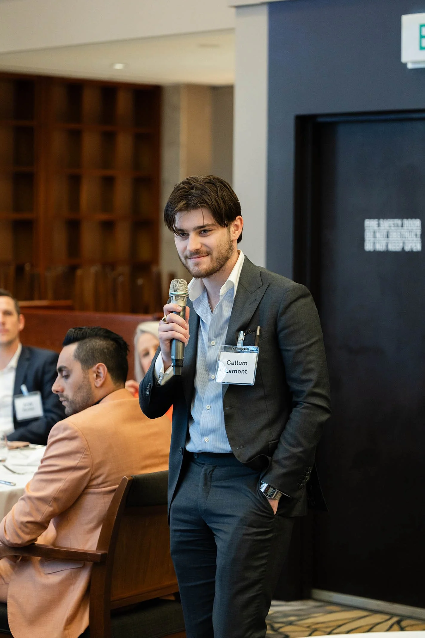 A young man with dark hair and a beard, wearing a suit and white shirt, stands with his hand in his pocket while holding a microphone in a conference room. He has a name tag that reads 'Callum Lamont' and is standing against a black wall near other s