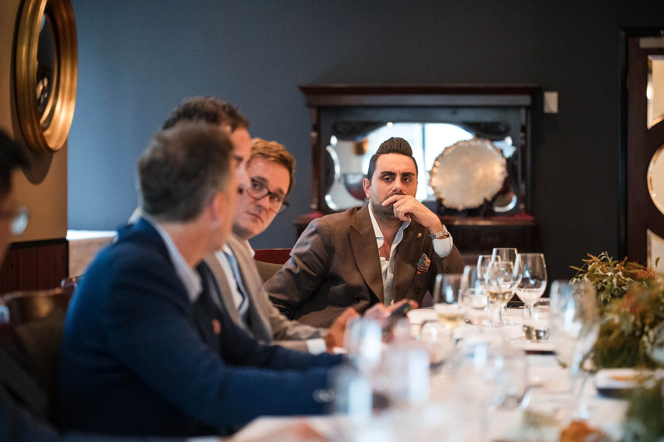 A group of men sitting at a formal dinner table, with one man in focus looking serious and thoughtful, while others are blurred in the foreground, in a dimly lit room with dark-colored walls and decorative items.