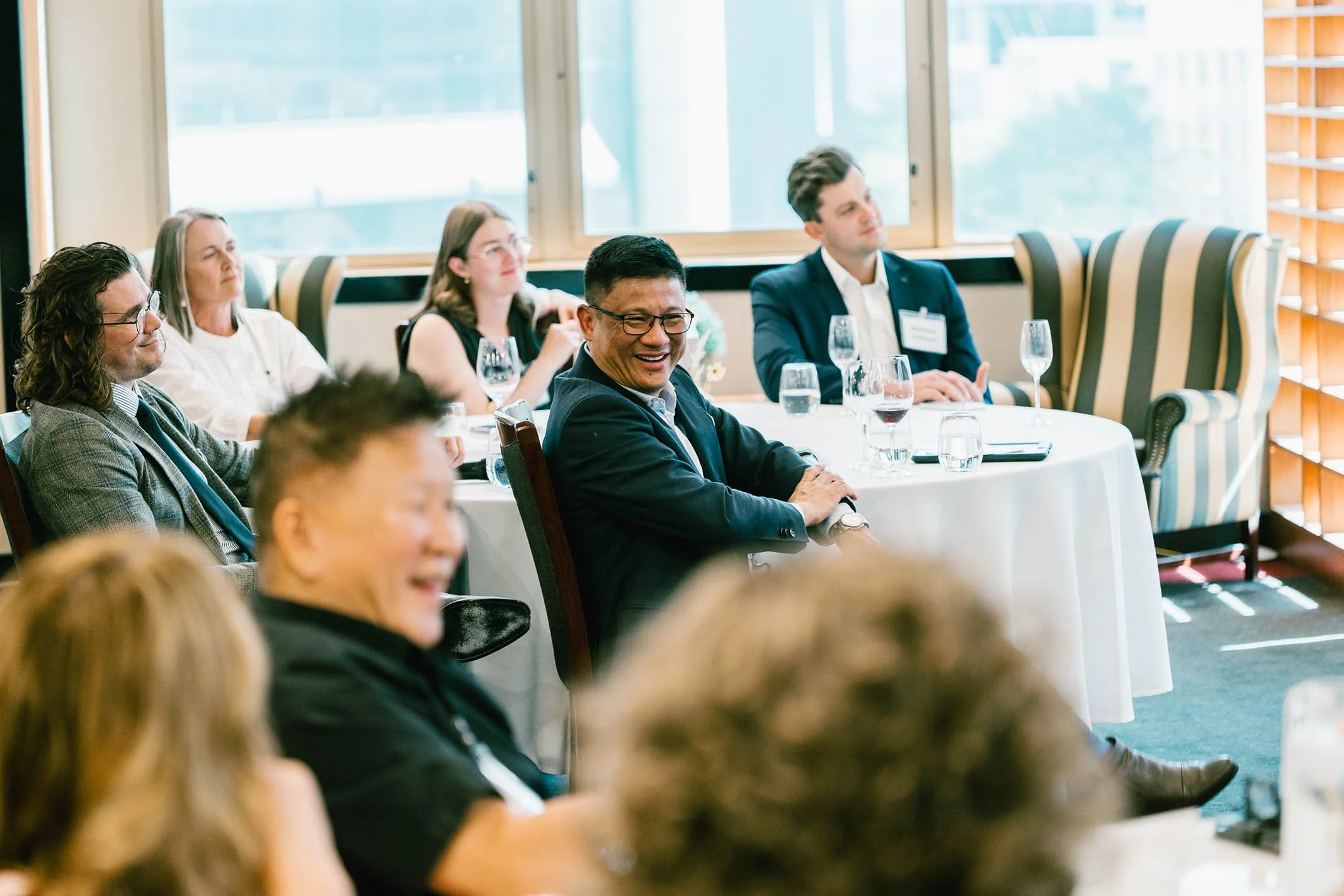 People sitting at a round table in a conference room, smiling and listening to a speaker, with some glasses of water on the table.