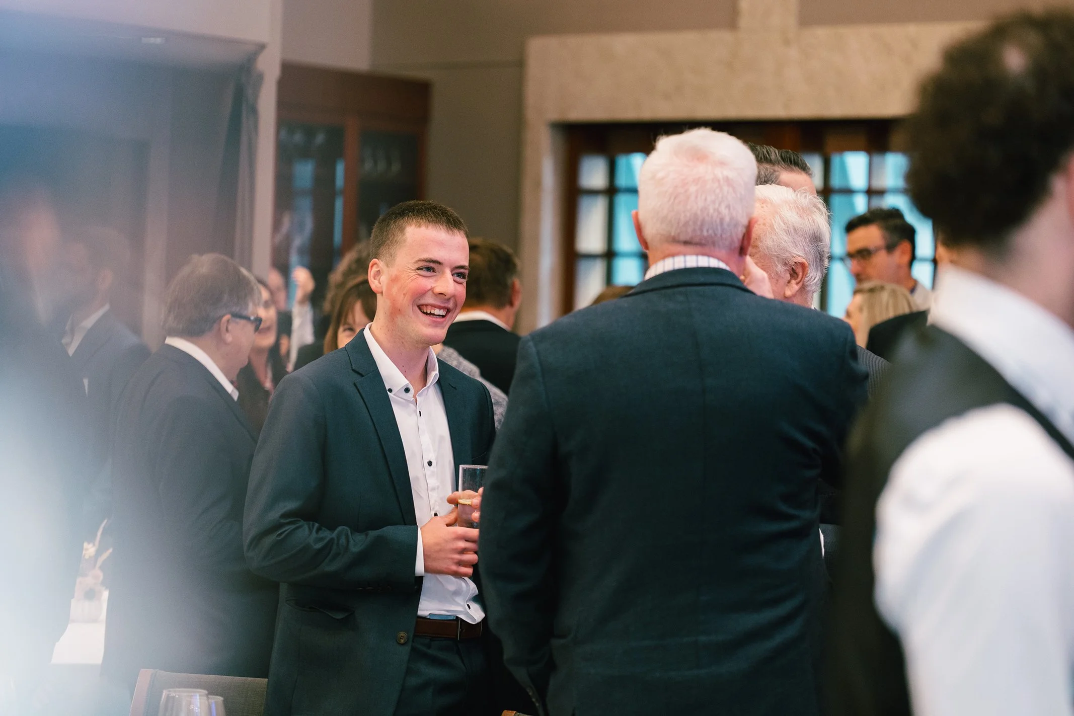 Group of people at a formal event, engaging in conversation. A young man with short dark hair, wearing a dark suit and white shirt, is smiling and holding a drink, standing near older individuals in suits. The setting appears to be indoors, well-lit 