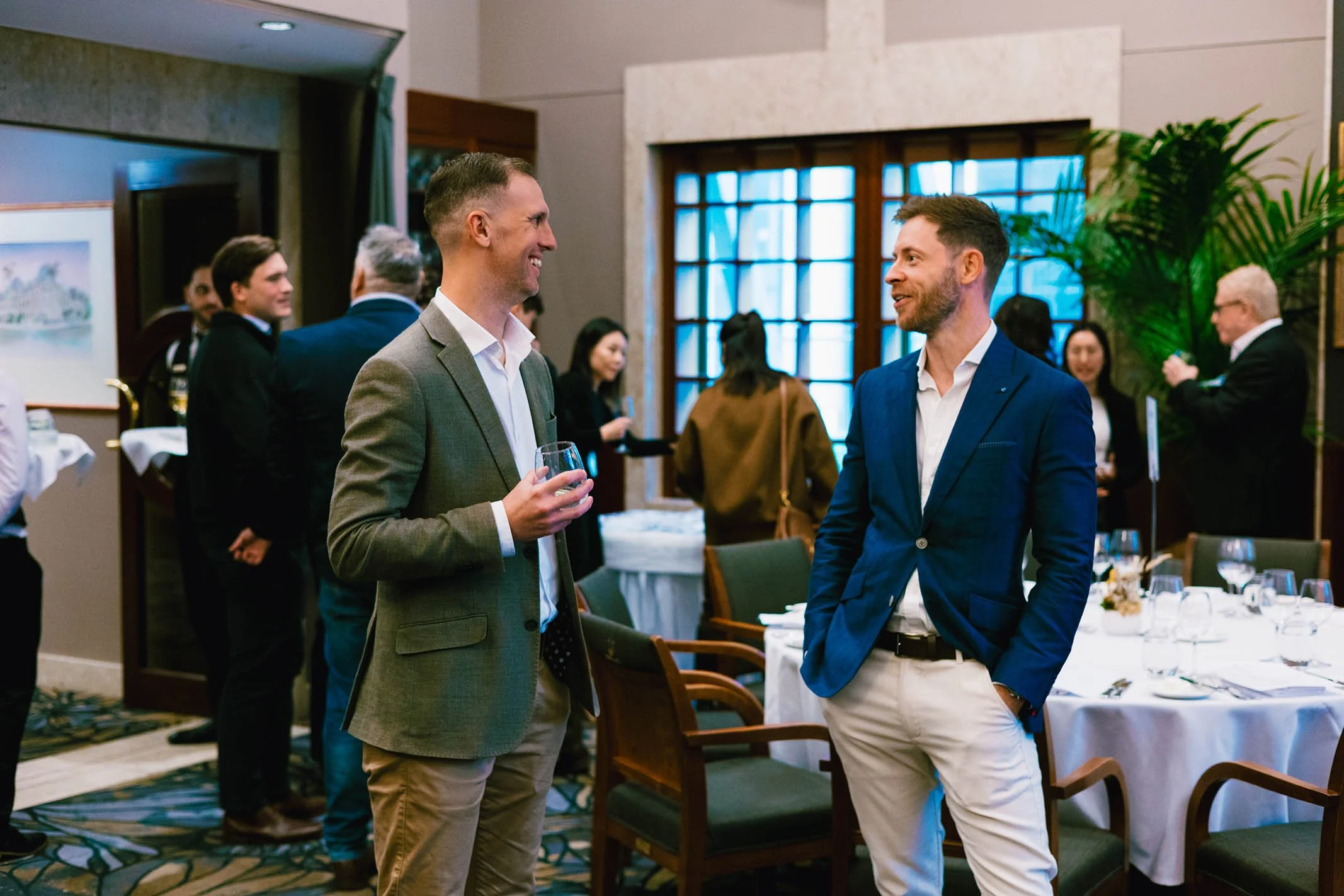 Two men in suits engaging in a conversation at a formal event with other guests in the background, dining tables, and large windows.