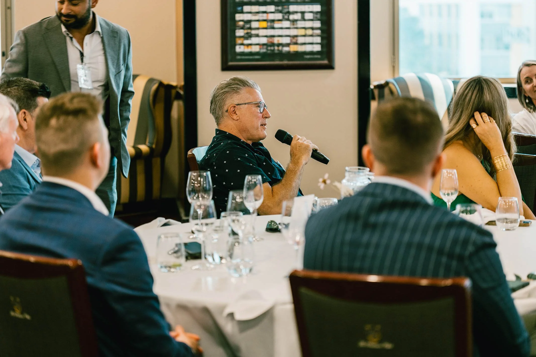 A man with gray hair and glasses is speaking into a microphone at a formal meeting or conference, seated at a round table covered with a white tablecloth, surrounded by other attendees dressed in business attire, with wine glasses and tableware, in a
