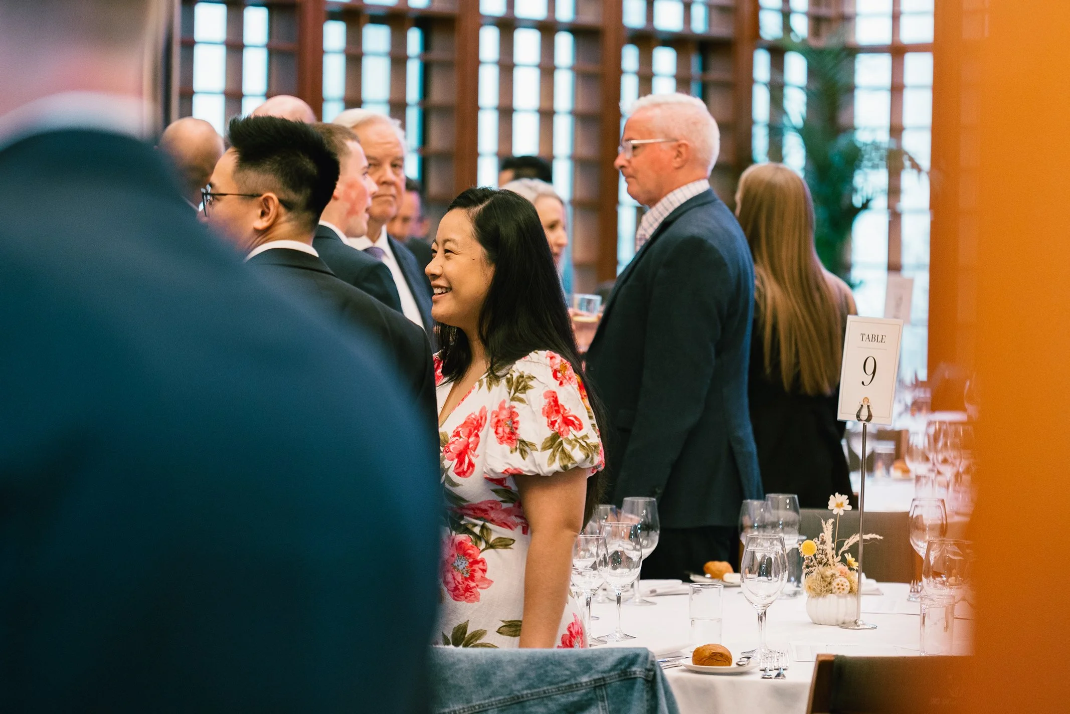 People gathered at a formal dining event, standing around tables with wine glasses and table settings, in a room with large windows and wooden decor.