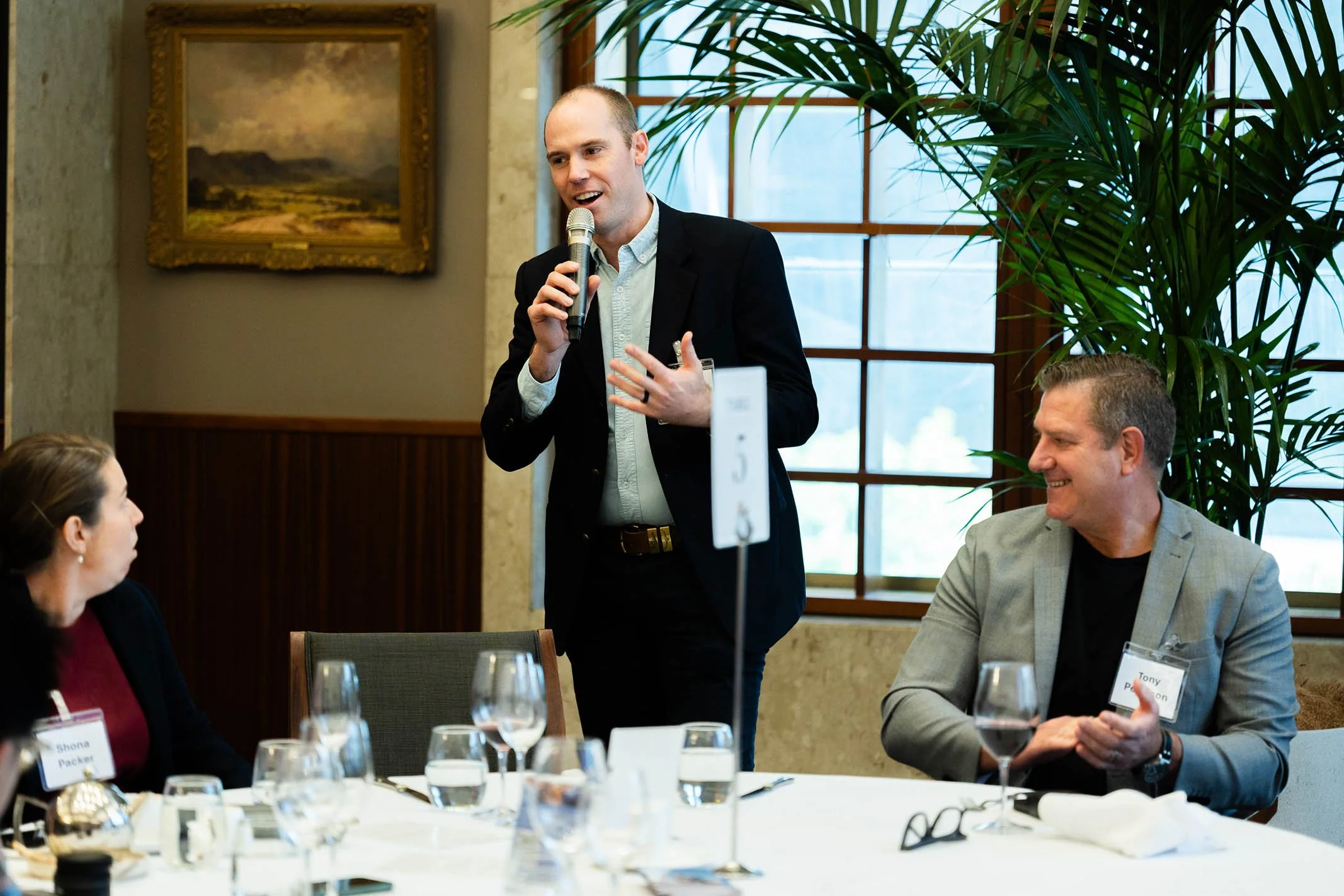 A man in a black blazer speaking into a microphone at a dinner event, seated at a table with two other people listening and smiling. The setting is a well-lit room with large windows, a plant, and framed artwork on the wall.