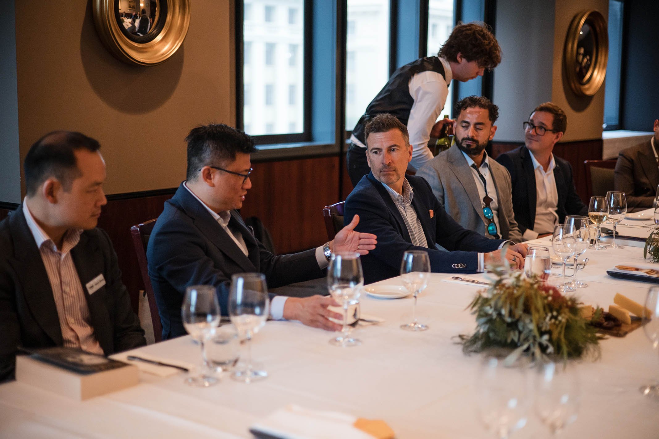 A group of people in formal business attire sitting at a long dining table during a conference or meeting, with a person standing behind them serving drinks.