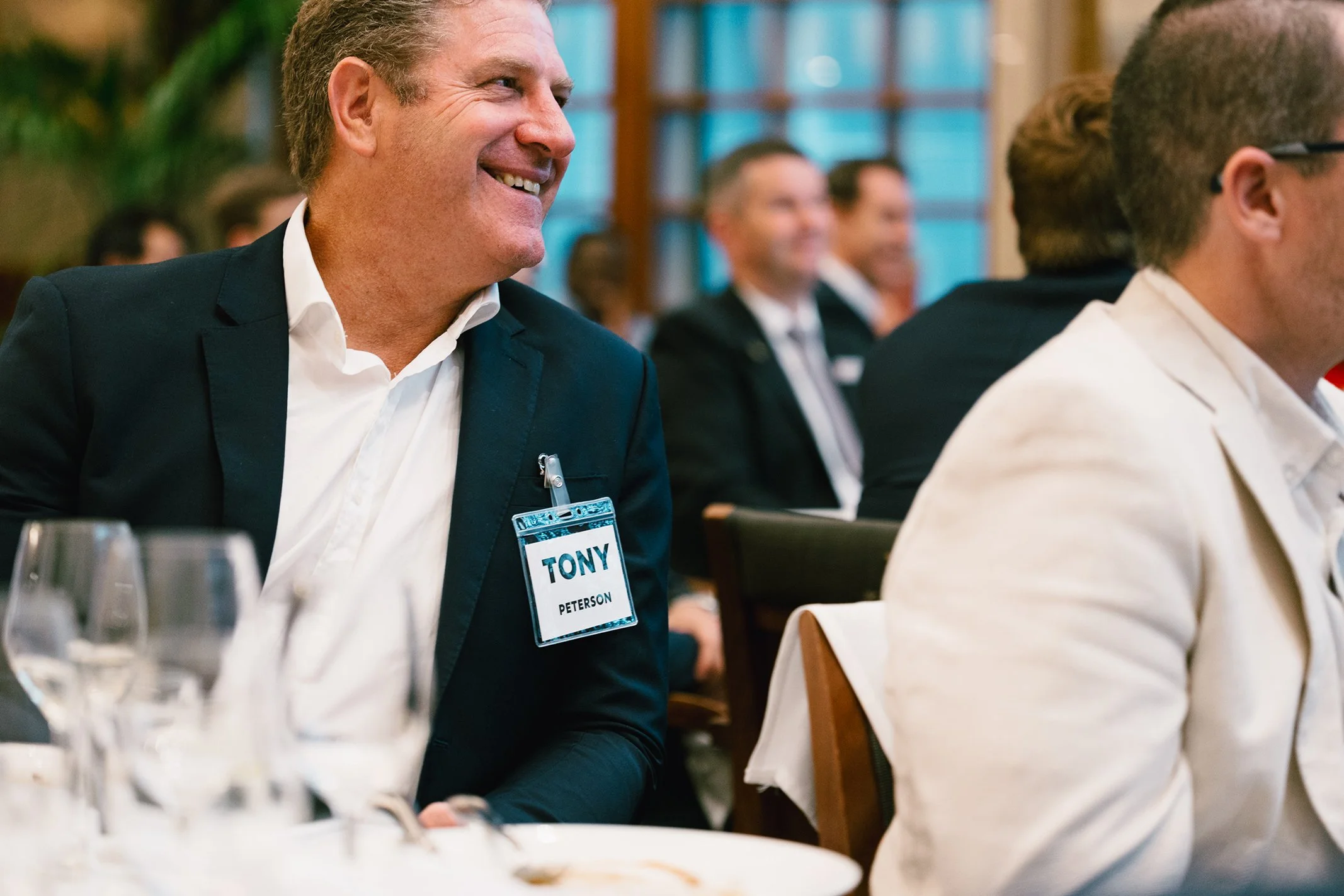 Man wearing a name tag that reads 'Tony Peterson' at a conference or meeting, smiling and sitting among other attendees.