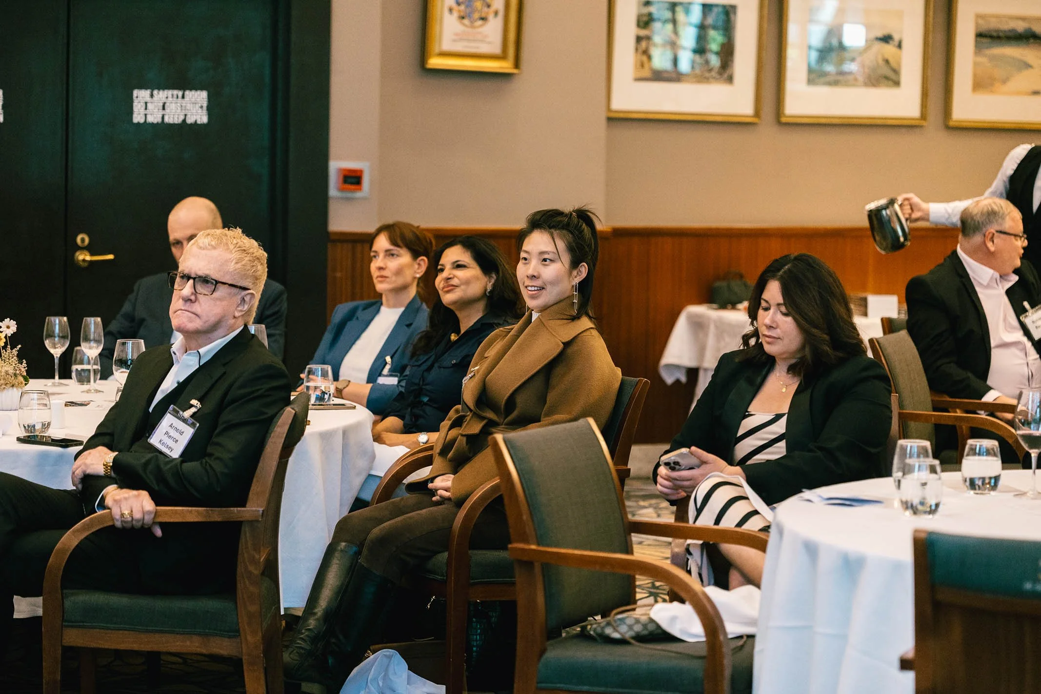 People sitting at a conference or meeting, listening attentively, with wine glasses on tables, in a wood-paneled room with framed artwork on the walls.