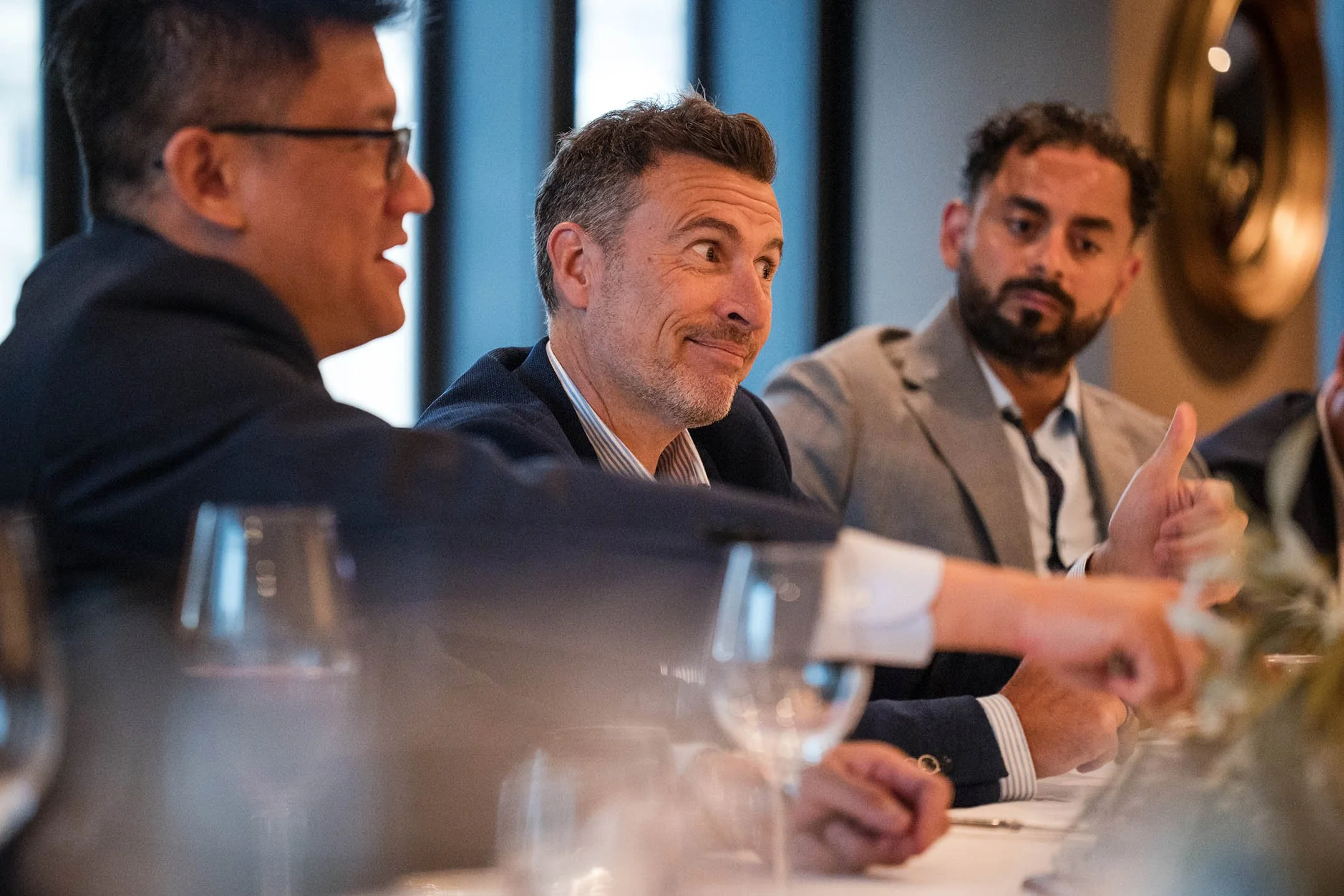 Three men in suits sitting at a table during a meeting or conference, with one men smiling and the third giving a thumbs up.