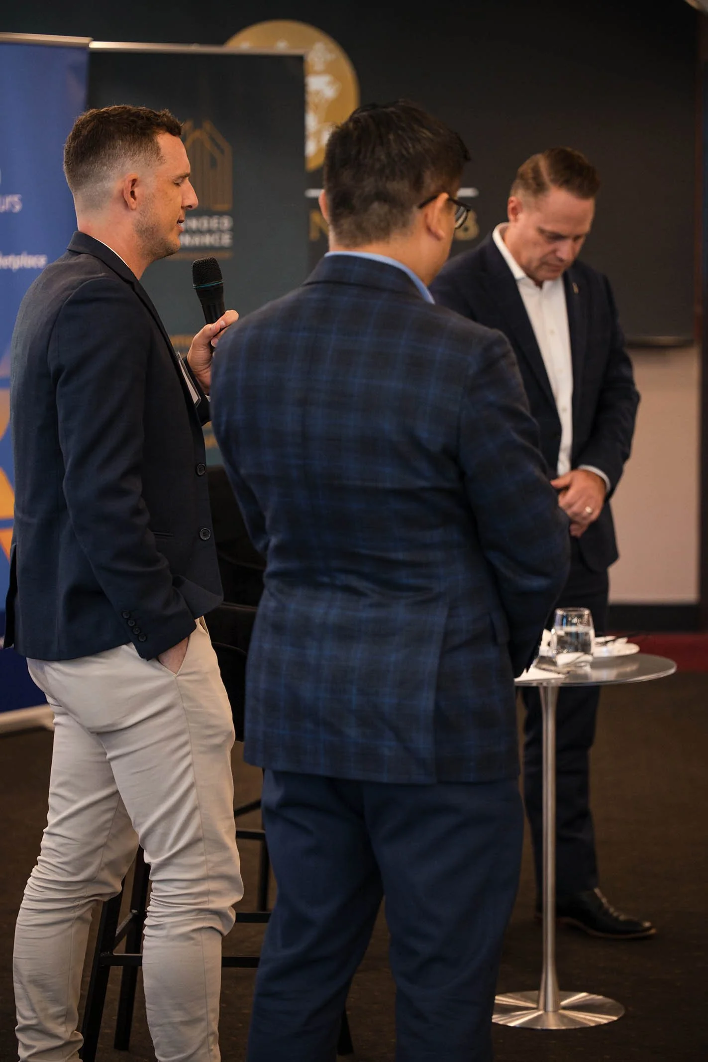 Three men in business attire standing around a small round table during a professional event. One man is speaking into a microphone, while the others listen with their heads bowed. The background shows a blue banner and a wall with a logo.