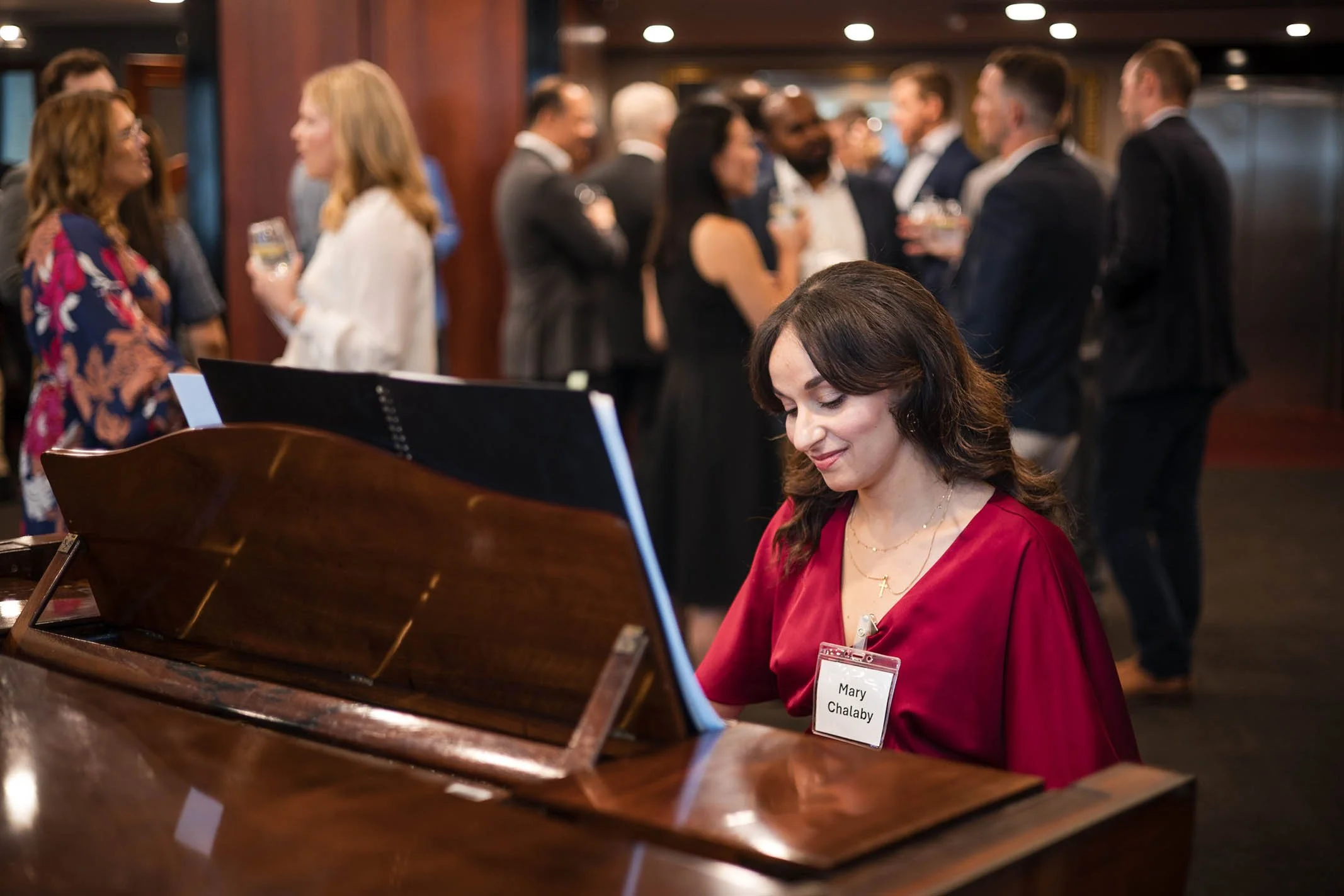 A woman in a red dress playing a piano at a social event or gathering, with a name tag that reads 'Mary Chalaby'. In the background, groups of people in formal attire are talking, holding drinks, and mingling in a room with warm lighting.