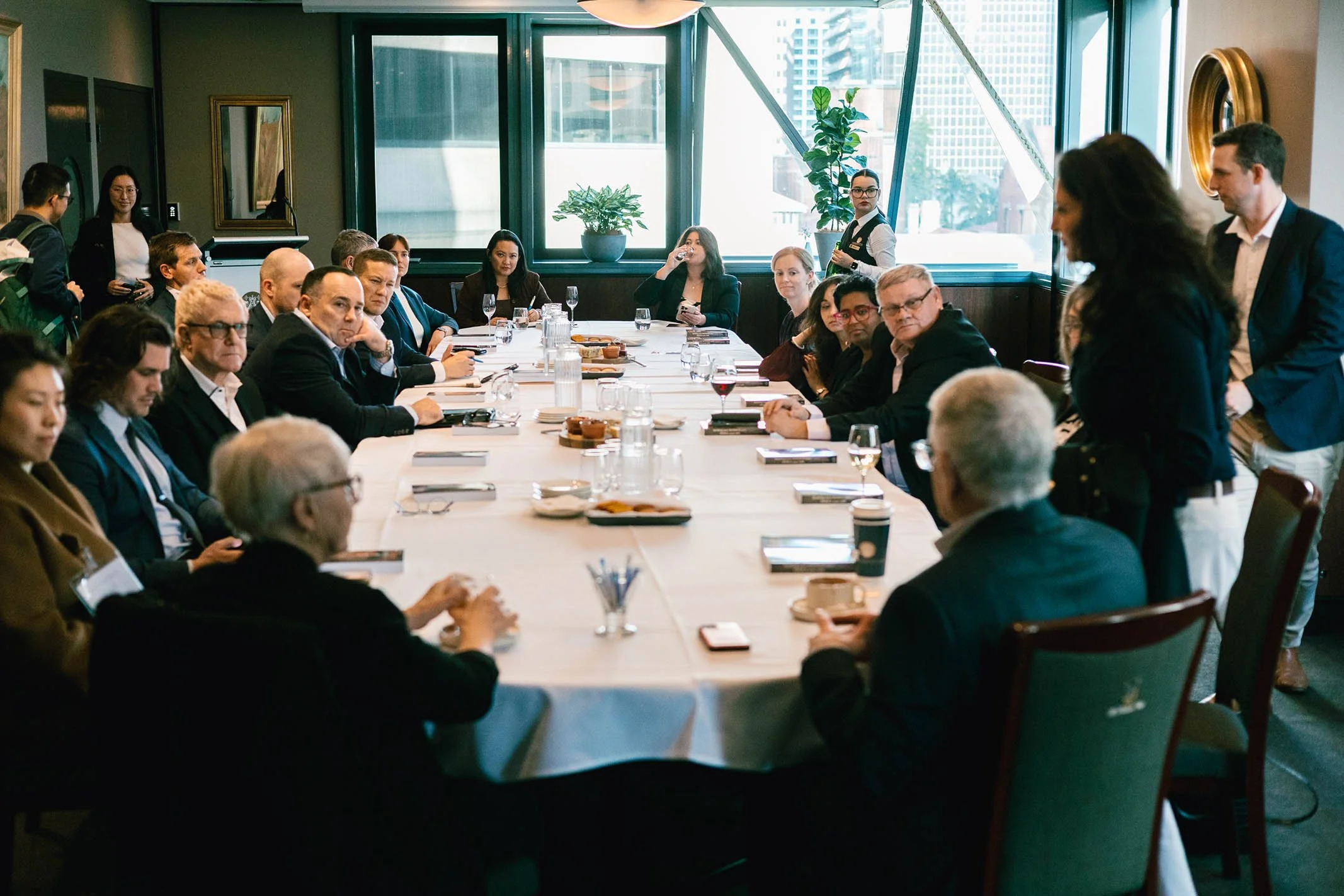 Business meeting with diverse group of professionals seated around a large conference table in a high-rise office, with some standing and engaging in discussion.