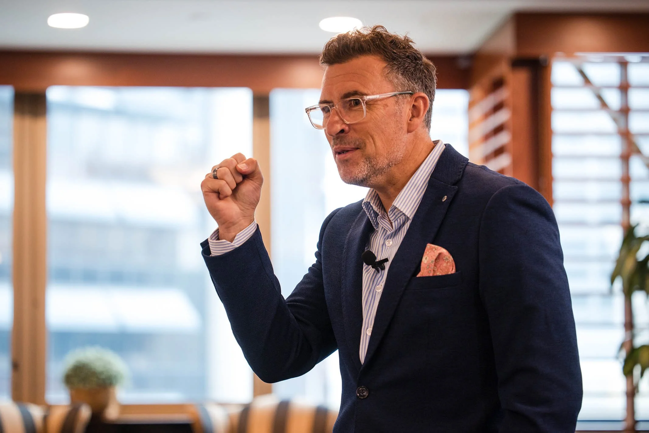 A middle-aged man wearing glasses and a navy blazer talks confidently in a modern office or conference room, gesturing with a raised fist.