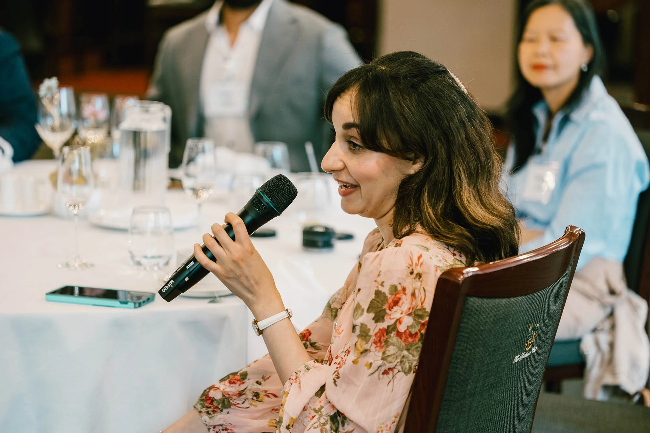 Woman speaking into a microphone at a formal event, sitting at a table with glassware and a smartphone, in a conference or banquet setting.