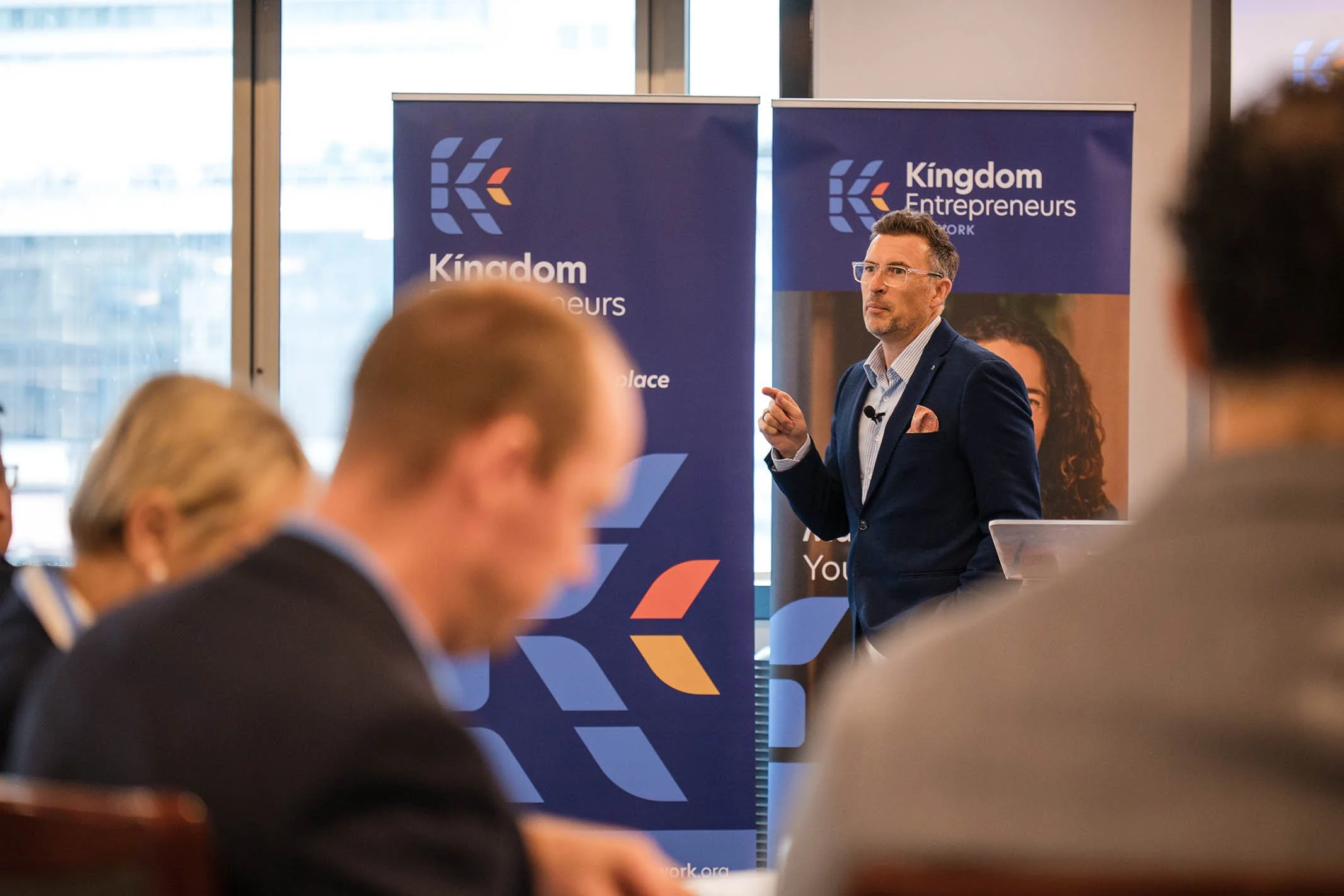 A man in a blue blazer and glasses giving a presentation at a conference, with a backdrop displaying the 'Kingdom Entrepreneurs Network' logo.