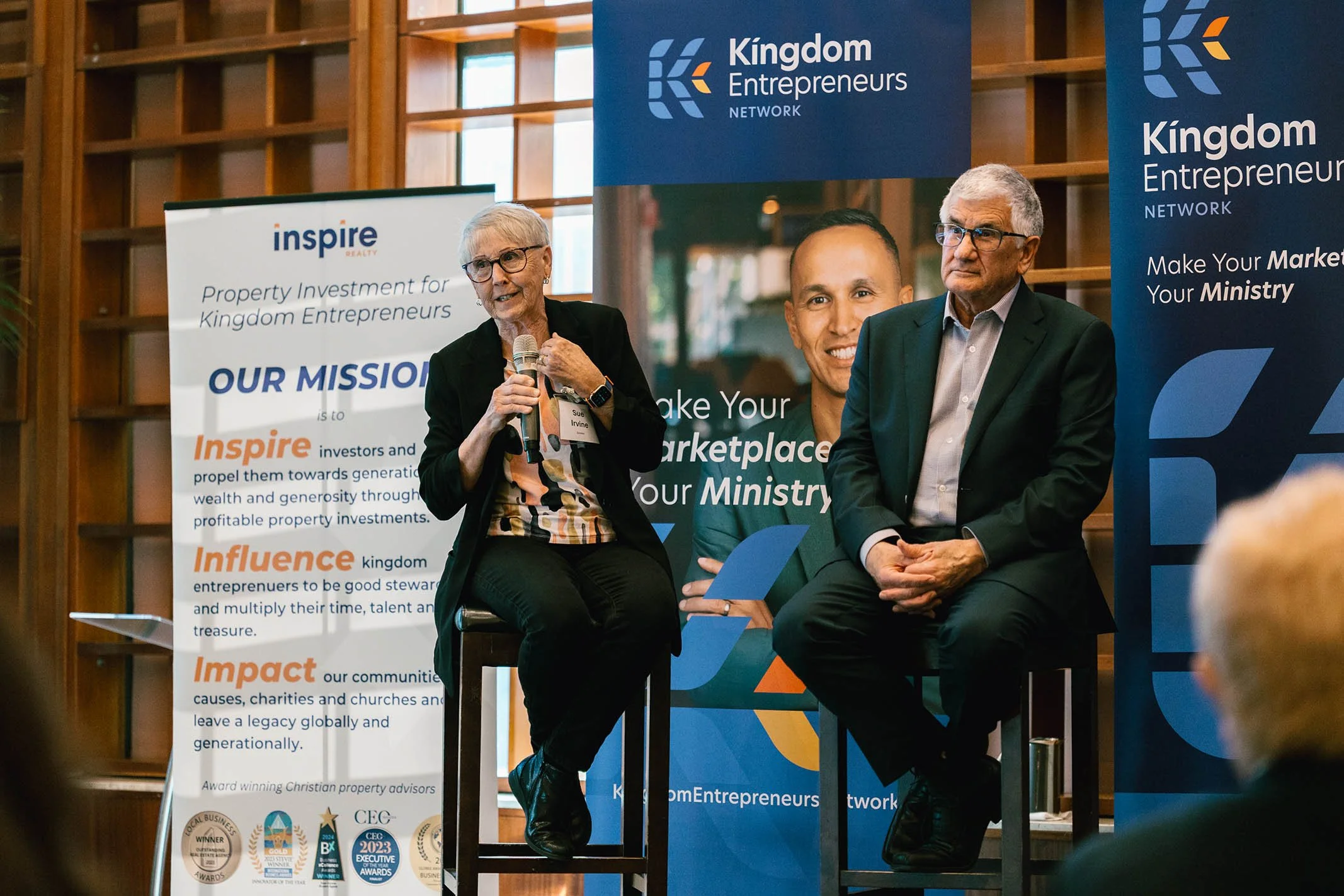 Two people, a woman and a man, sitting on chairs on a stage at a Kingdom Entrepreneurs Network event. The woman is speaking into a microphone, wearing a black blazer and glasses. The man is listening, dressed in a dark suit and glasses. Behind them a
