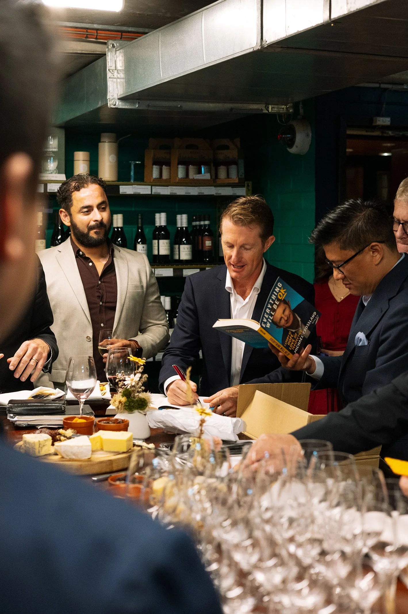 People gathered around a table at a social event, with wine bottles, cheese, glasses, and small flowers on the table, and shelves of wine bottles in the background.