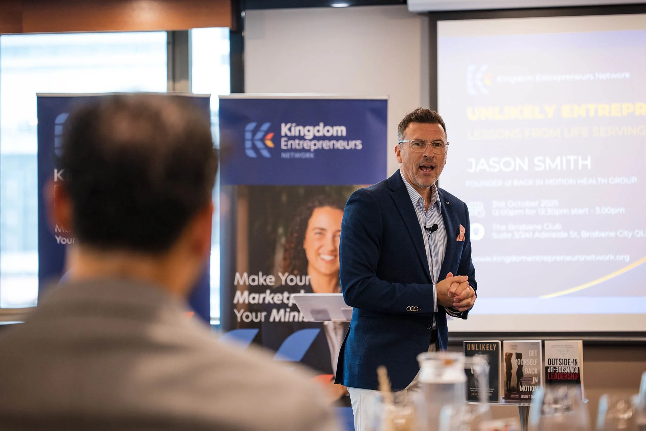 A man in a blue blazer giving a presentation at an event, with a Kingdom Entrepreneurs banner and a screen behind him displaying the title 'Unlikely Entrepreneurs' and the name Jason Smith.
