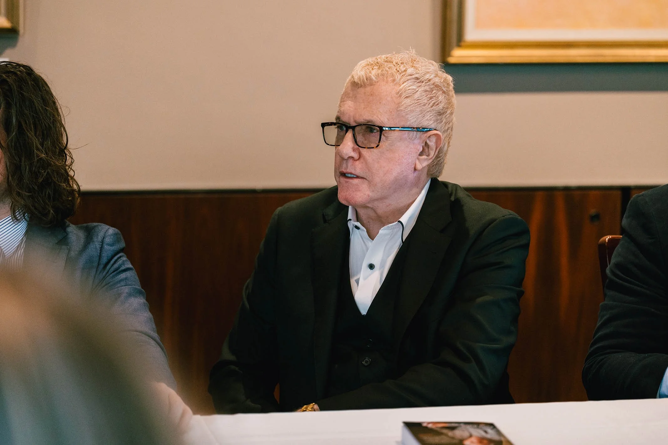 A man with light-colored hair, glasses, and a black suit is seated at a table, speaking during a meeting or conference.