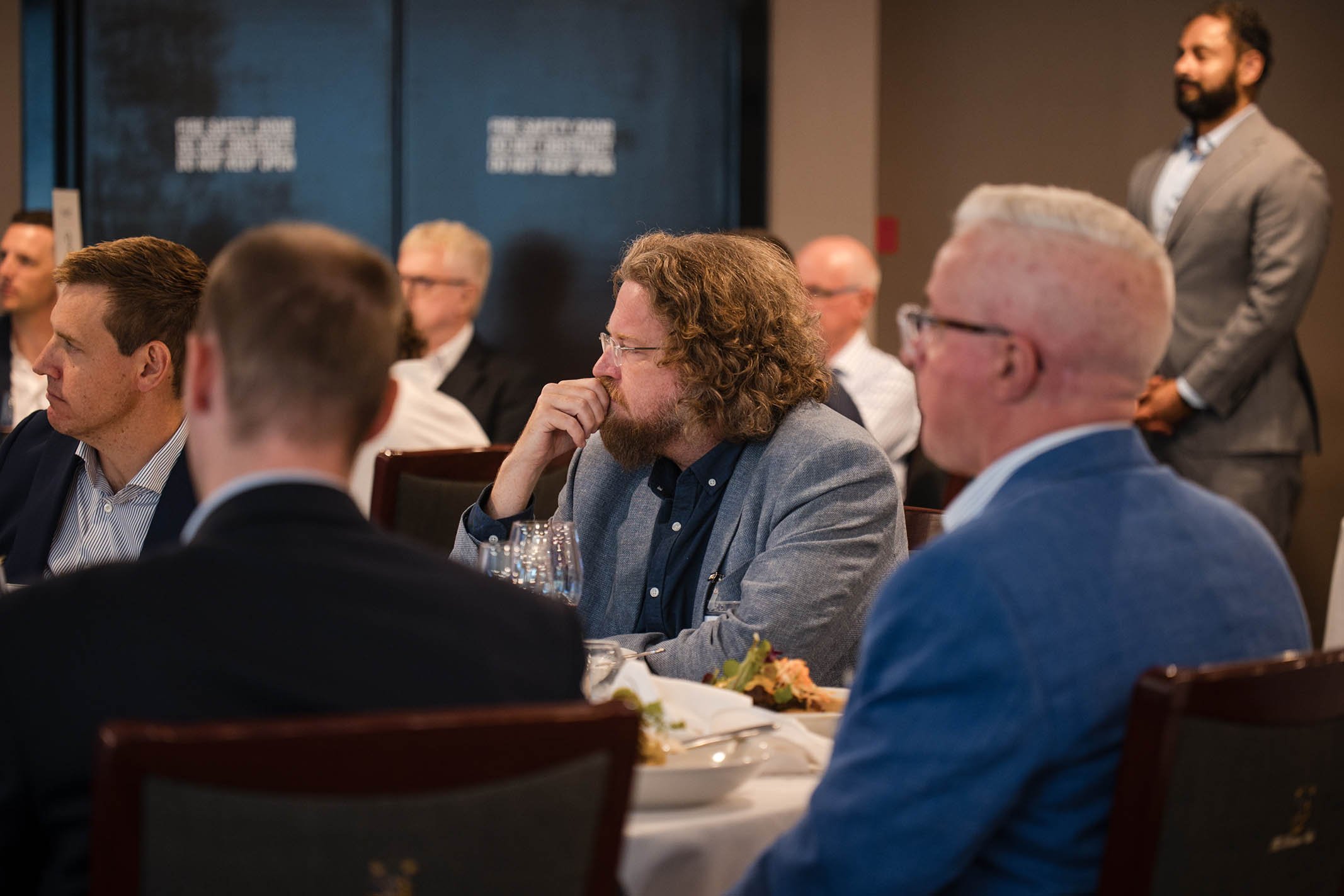 People attending a conference or business meeting, seated at a table with food and drinks, listening to a speaker or presentation in a formal setting.