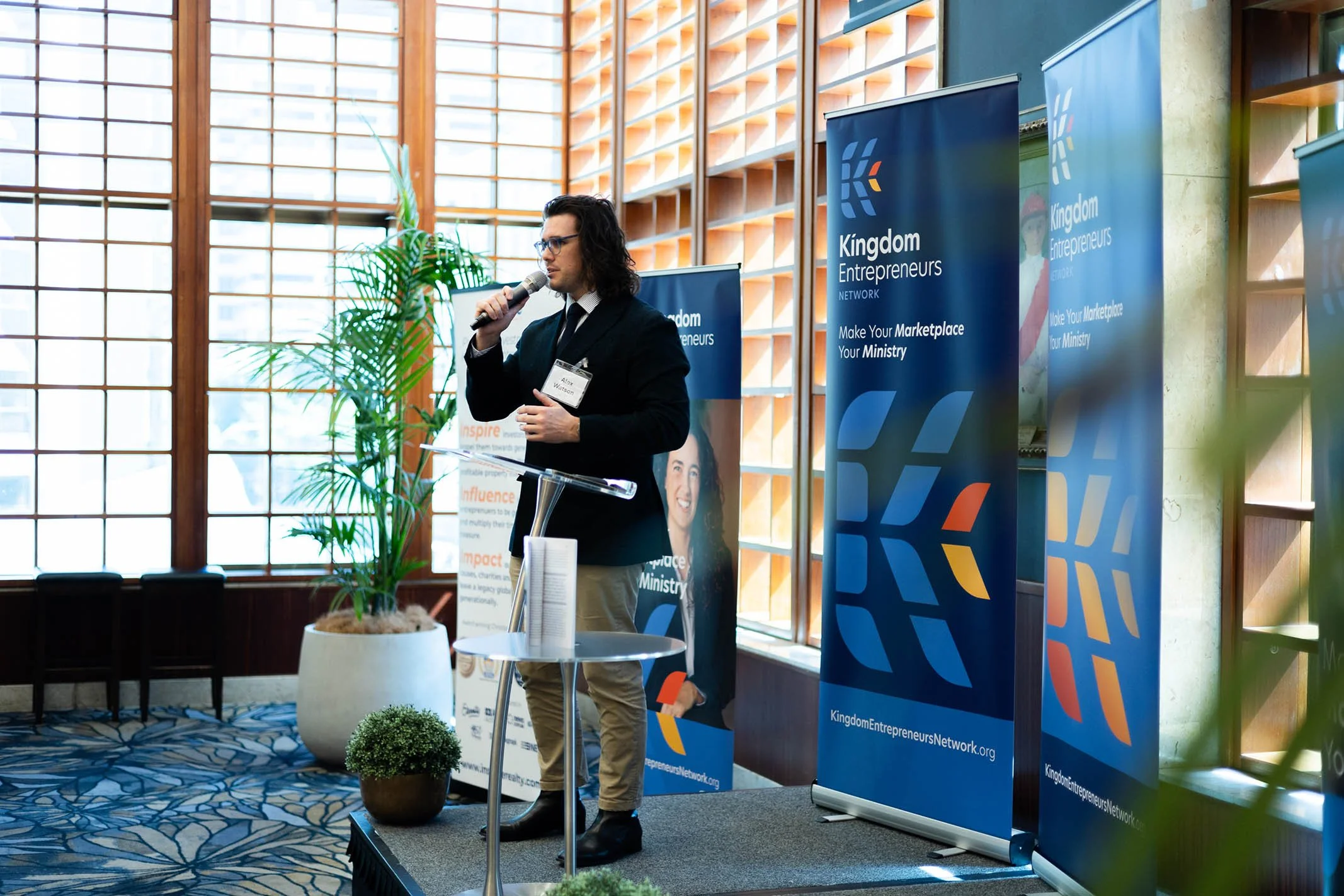 A man in a dark suit and glasses speaking into a microphone at a conference, standing behind a clear podium, with banners displaying 'Kingdom Entrepreneurs' in the background, in a room with large windows and indoor plants.