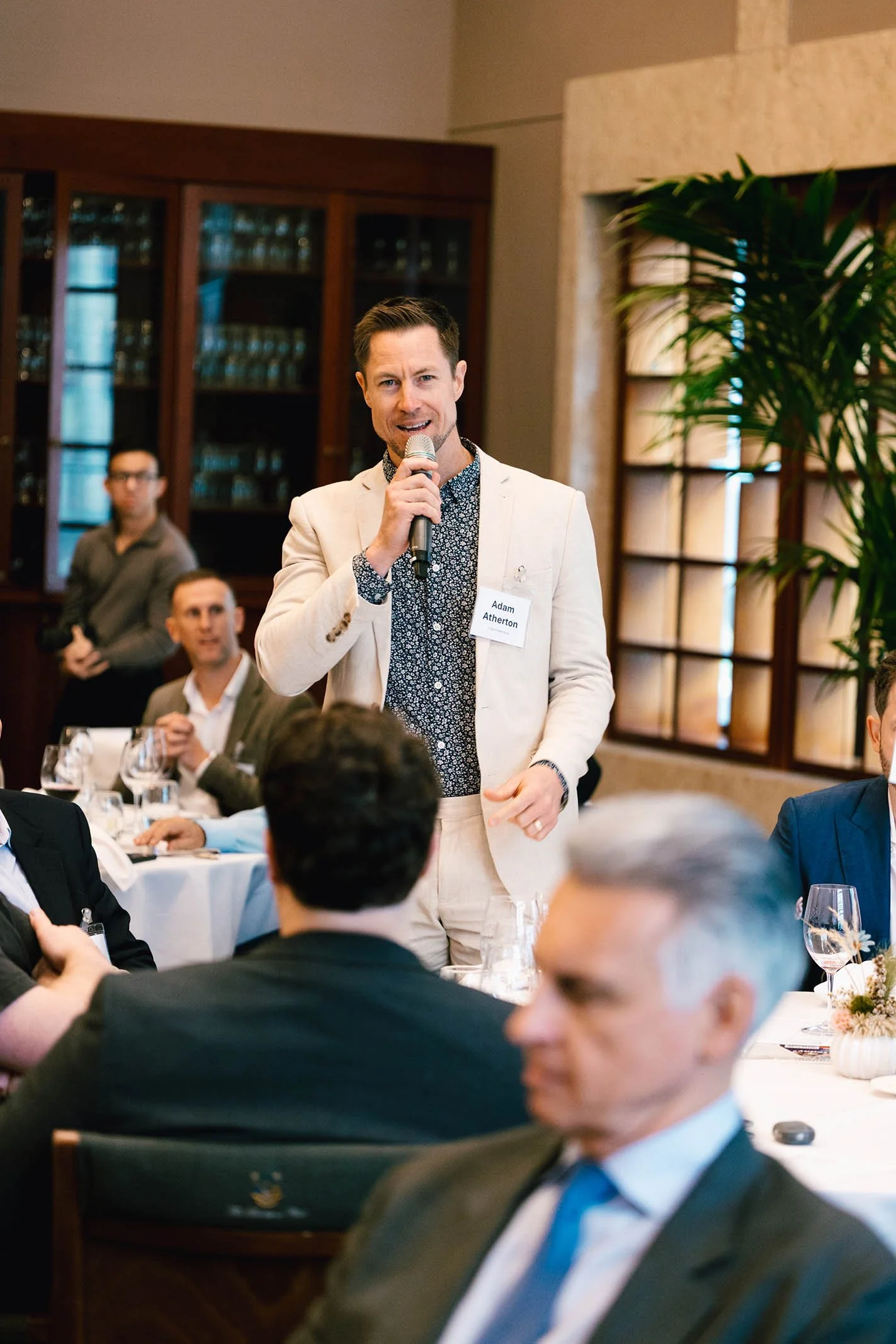 A man standing and speaking into a microphone at a formal event, wearing a beige suit and patterned shirt, with other attendees seated at round tables nearby.