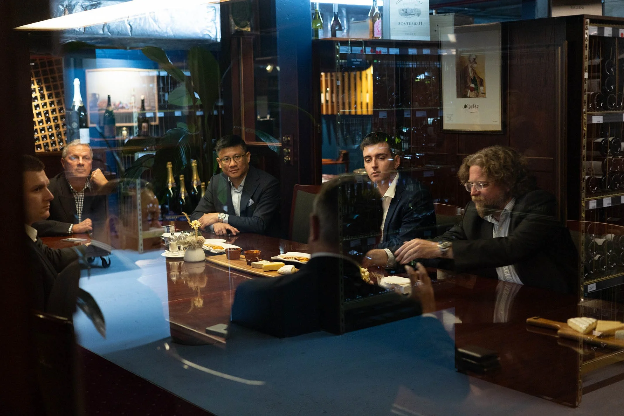 Group of six men discussing around a wine-tasting table in a wine cellar or restaurant, with bottles of wine and cheese on the table, seen through a glass window.