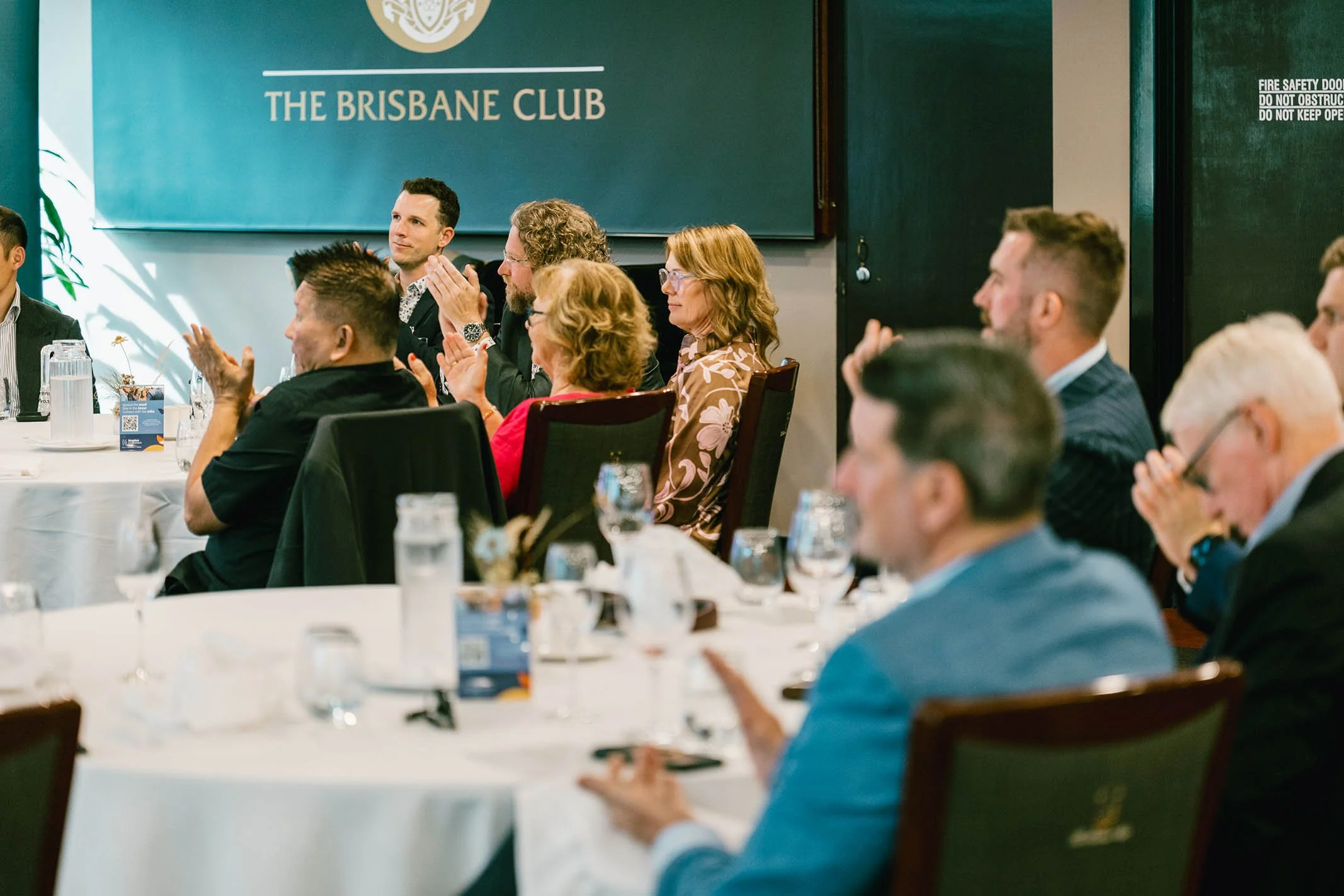 Group of people attending a formal event or conference at The Brisbane Club, sitting around round tables with glasses and bottles, some clapping and listening attentively.