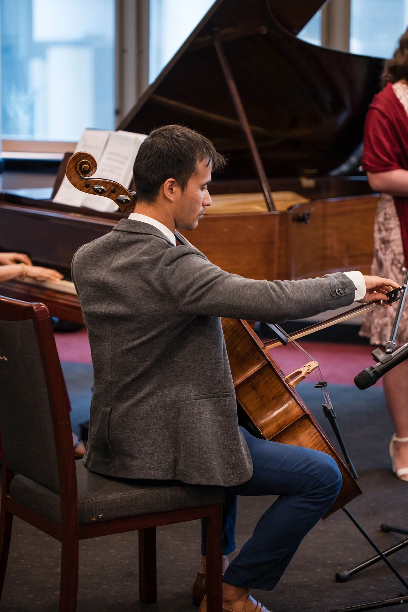 A man in a gray blazer playing a cello at a music performance, with a grand piano and a woman in a red dress nearby.