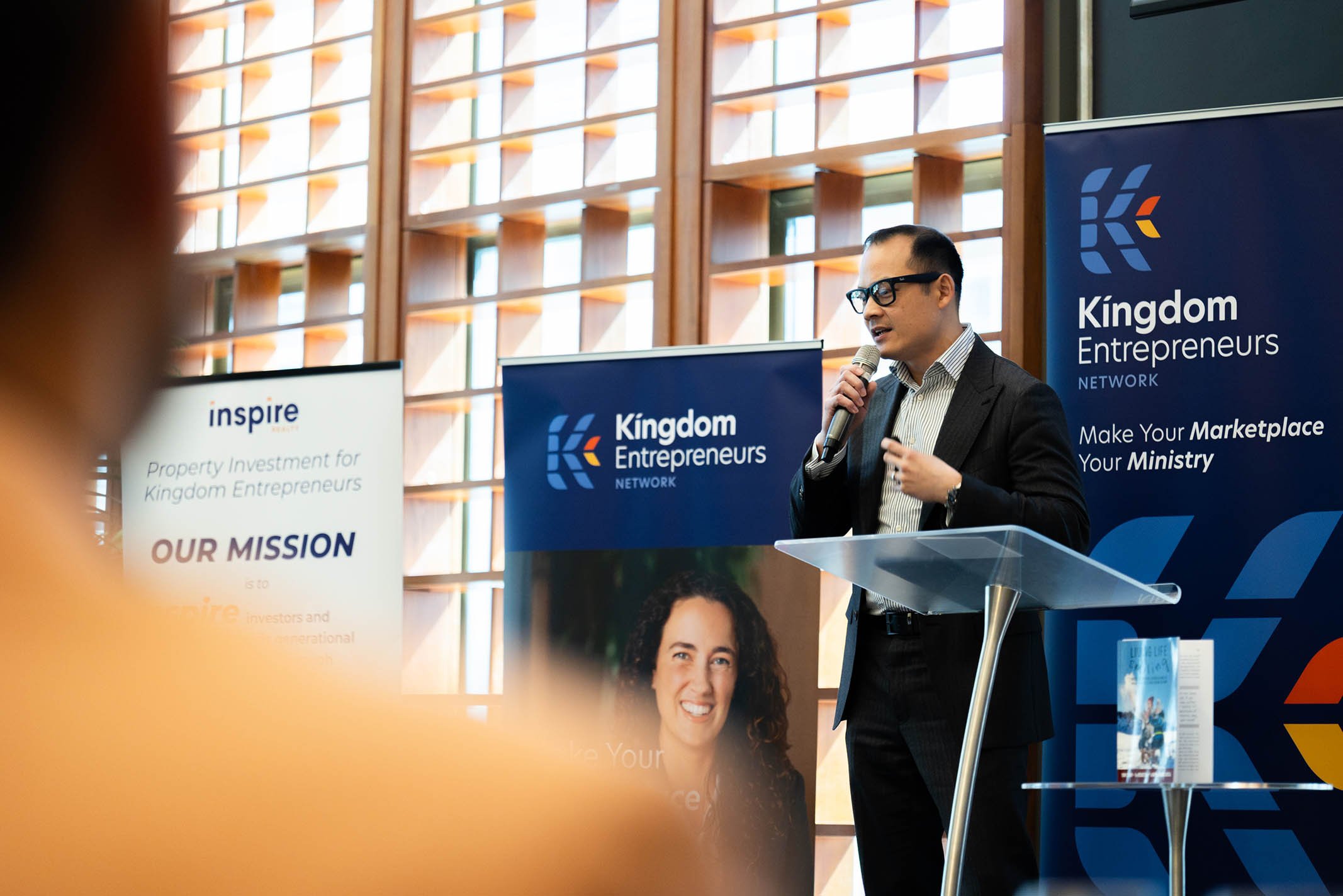 A man wearing glasses in a suit speaking into a microphone at a podium during a conference, with banners for Kingdom Entrepreneurs Network behind him and a partially visible banner with the word "inspire".