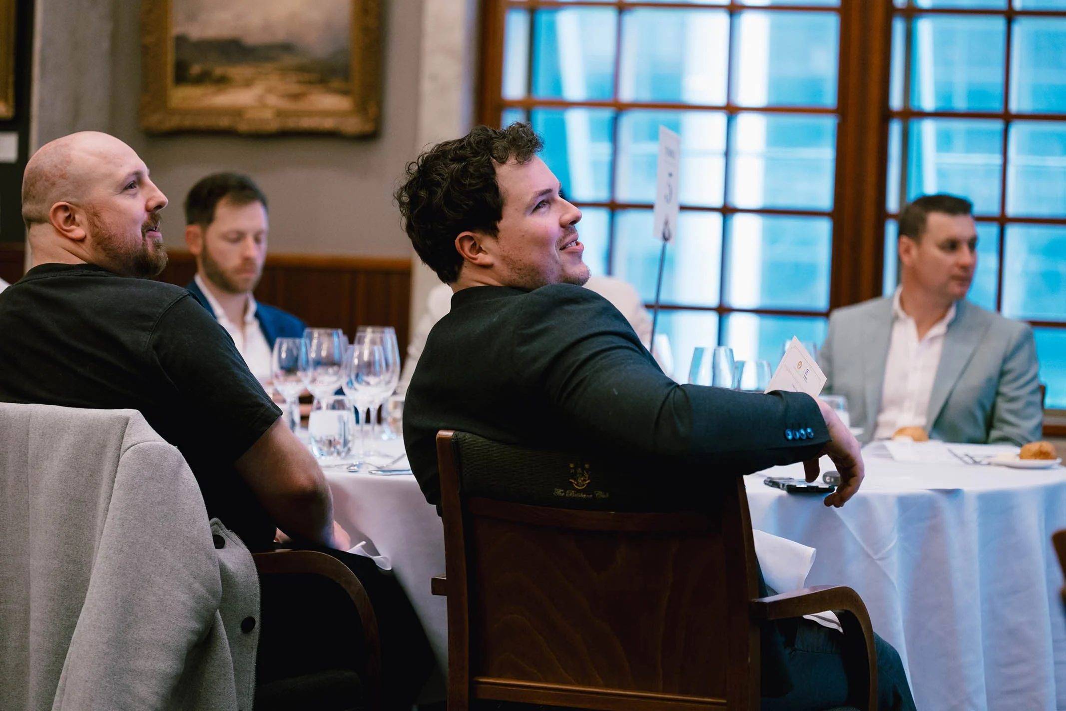 People sitting at a formal event table, with glasses and menus, inside a room with large windows and wooden paneling.
