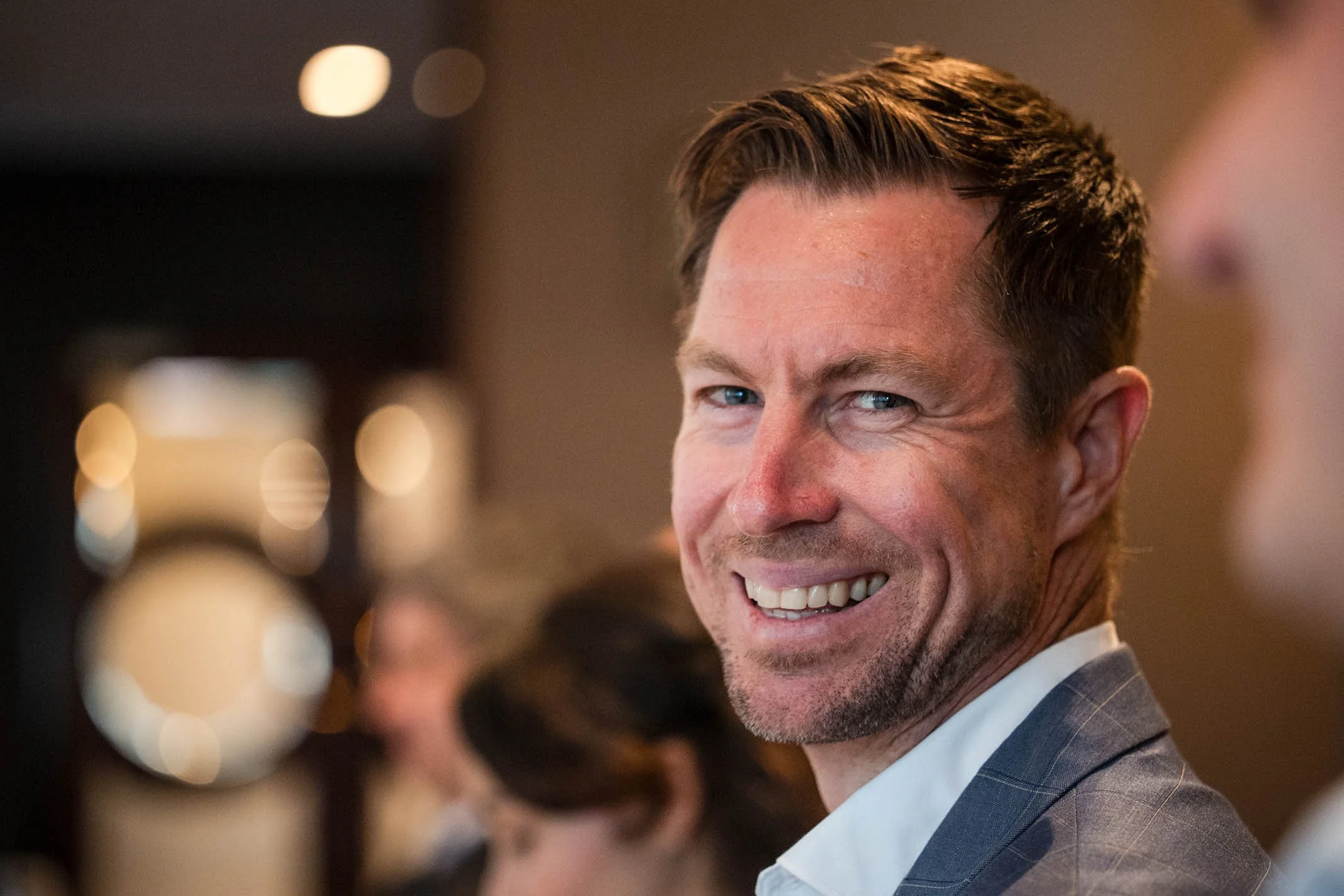 A smiling man with blue eyes and short brown hair, wearing a gray suit and white shirt, at a social event with blurred background lighting.