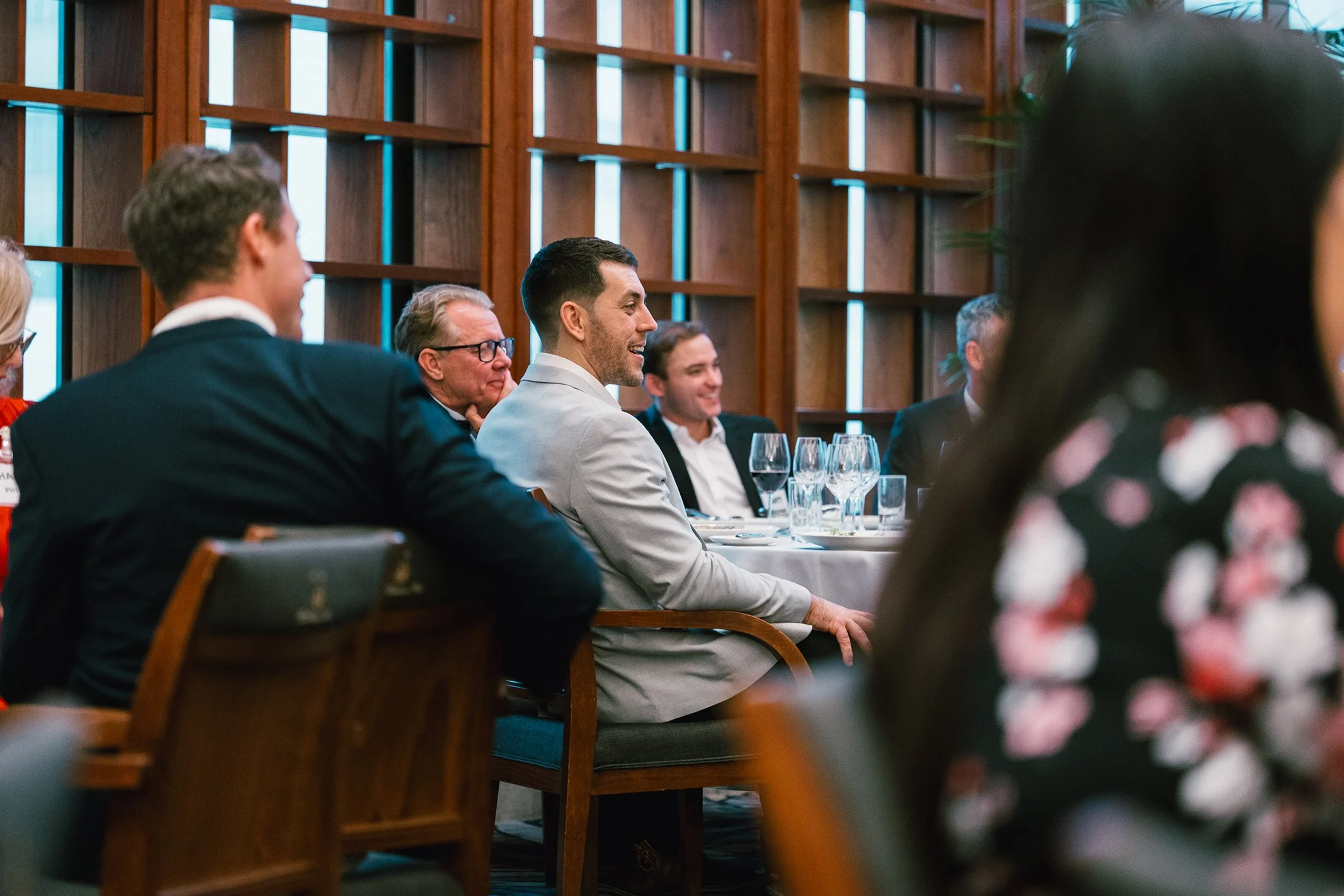 People attending a formal event seated at tables with wine glasses and tableware in a room with wooden paneling and large windows.