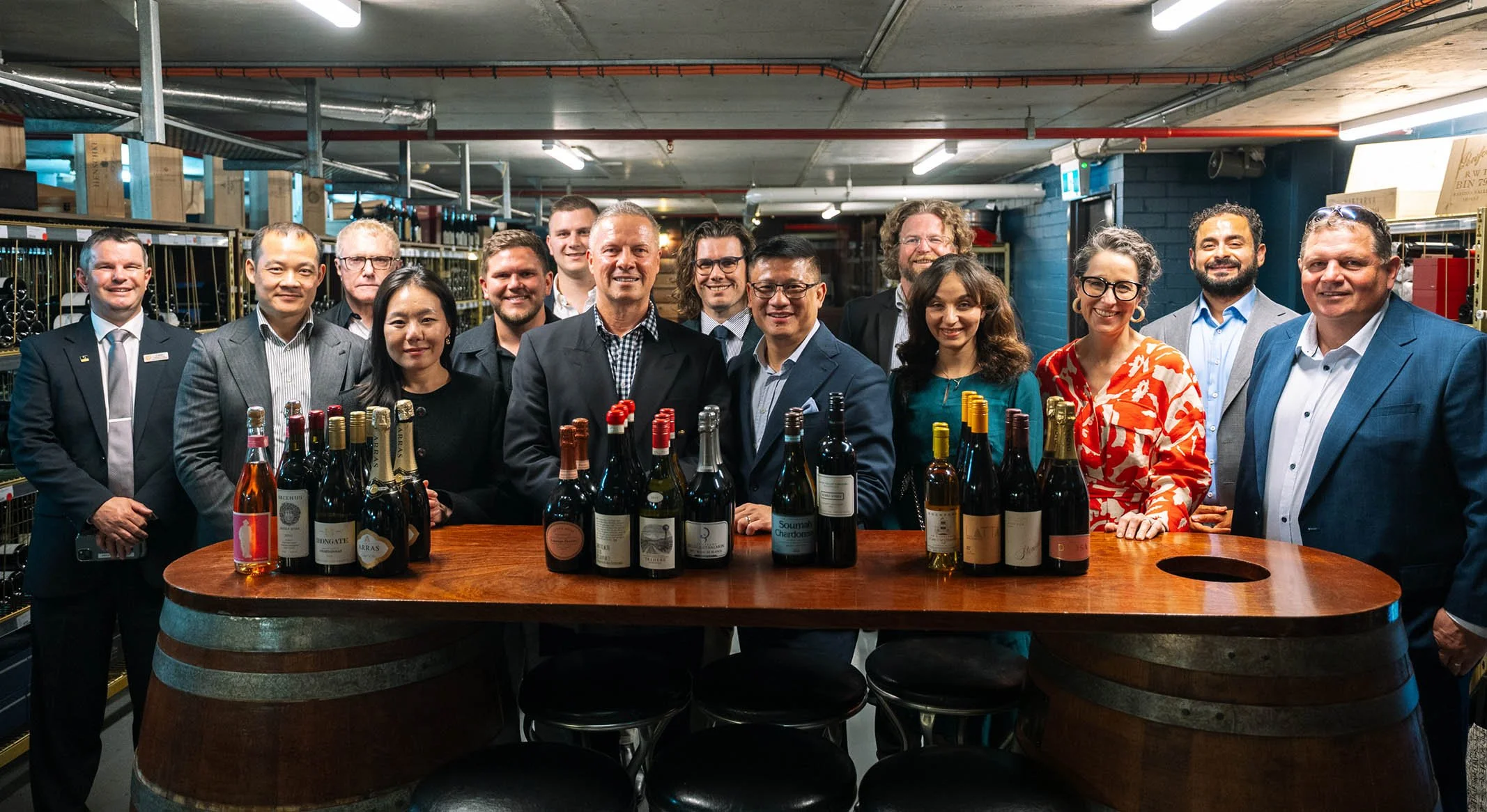 Group of people in formal attire gathered around a wooden table with wine bottles in a storage or warehouse setting.