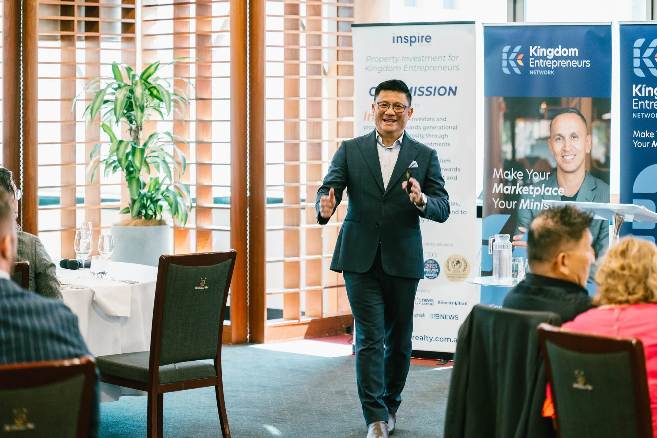 A man in a dark suit and glasses is giving a presentation in a conference room, standing near promotional banners for Kingdom Entrepreneurs and Inspire with a group of seated attendees.