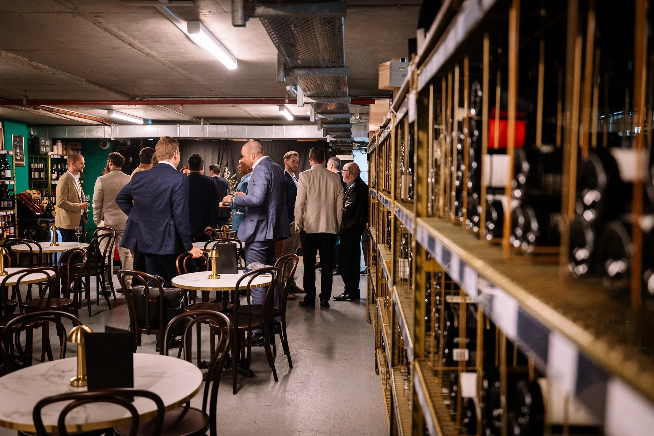 Group of men in suits socializing at a bar or restaurant, with wine bottles on shelves to the right and empty tables with chairs in the foreground.