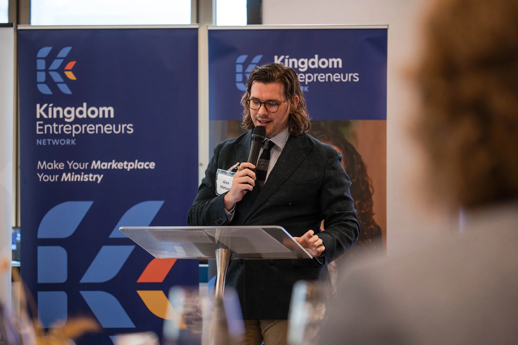 A man with long hair and glasses speaking into a microphone at a podium during an event with Kingdom Entrepreneurs banners in the background.