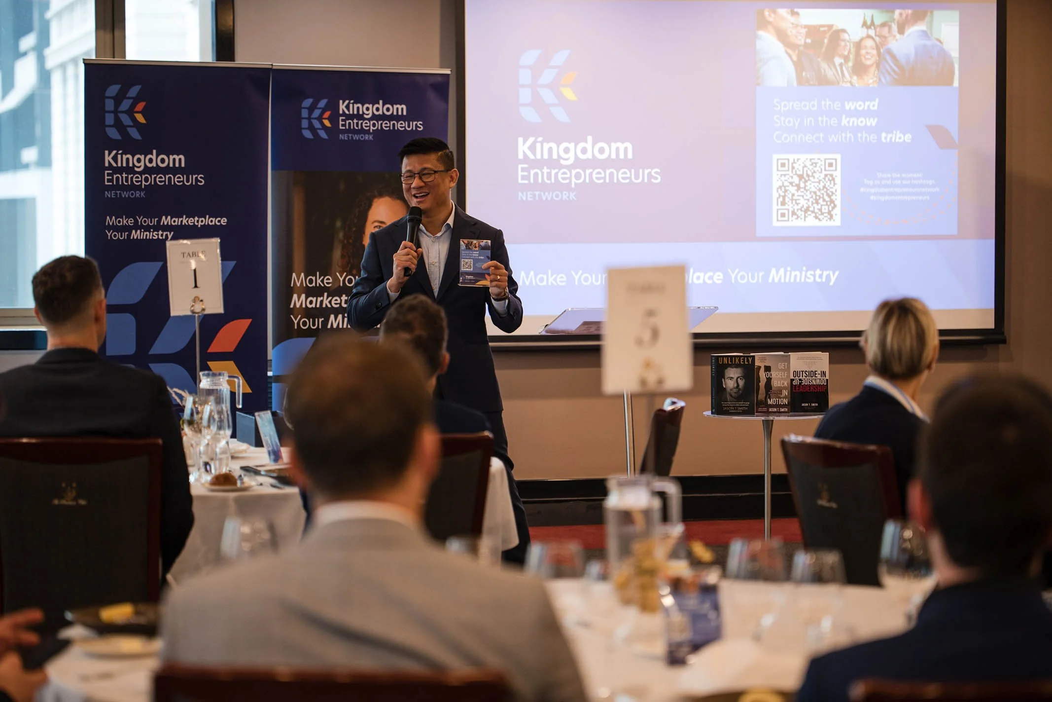 A man in a dark suit holding a microphone and a pamphlet, standing in front of a screen and banners at a professional event, with an audience seated at tables in a conference room.