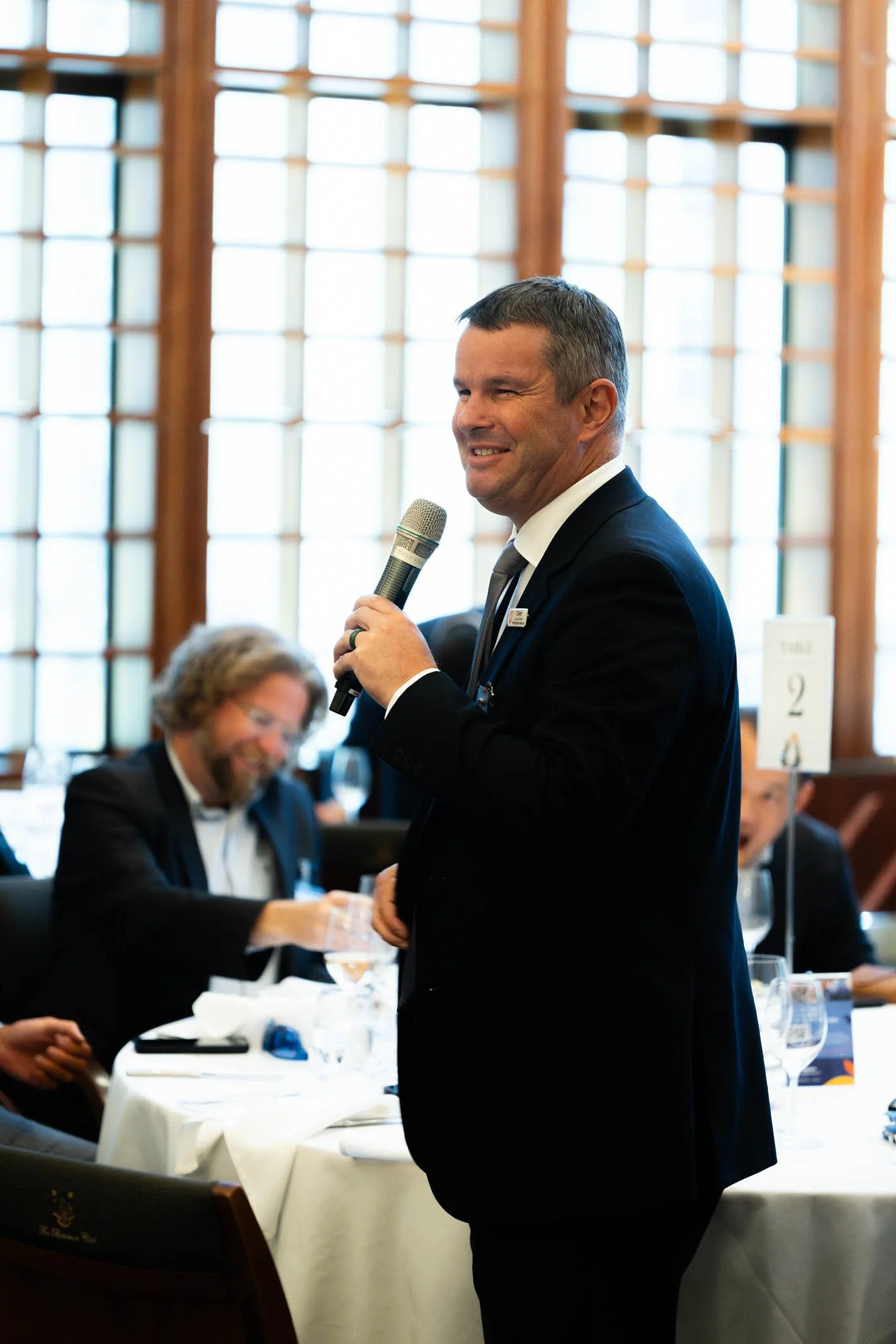 A man in a dark suit holding a microphone while speaking at a formal event with several seated people in the background, in a room with large wooden windows.